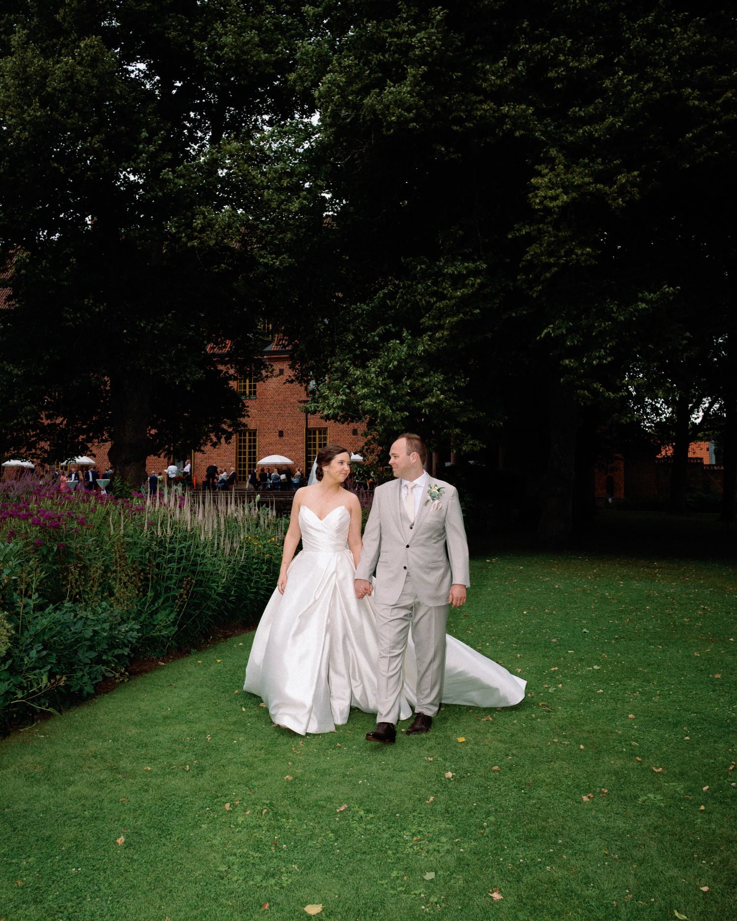 Bride and groom walk hand in hand through lush garden grounds with red brick building backdrop on their wedding day.