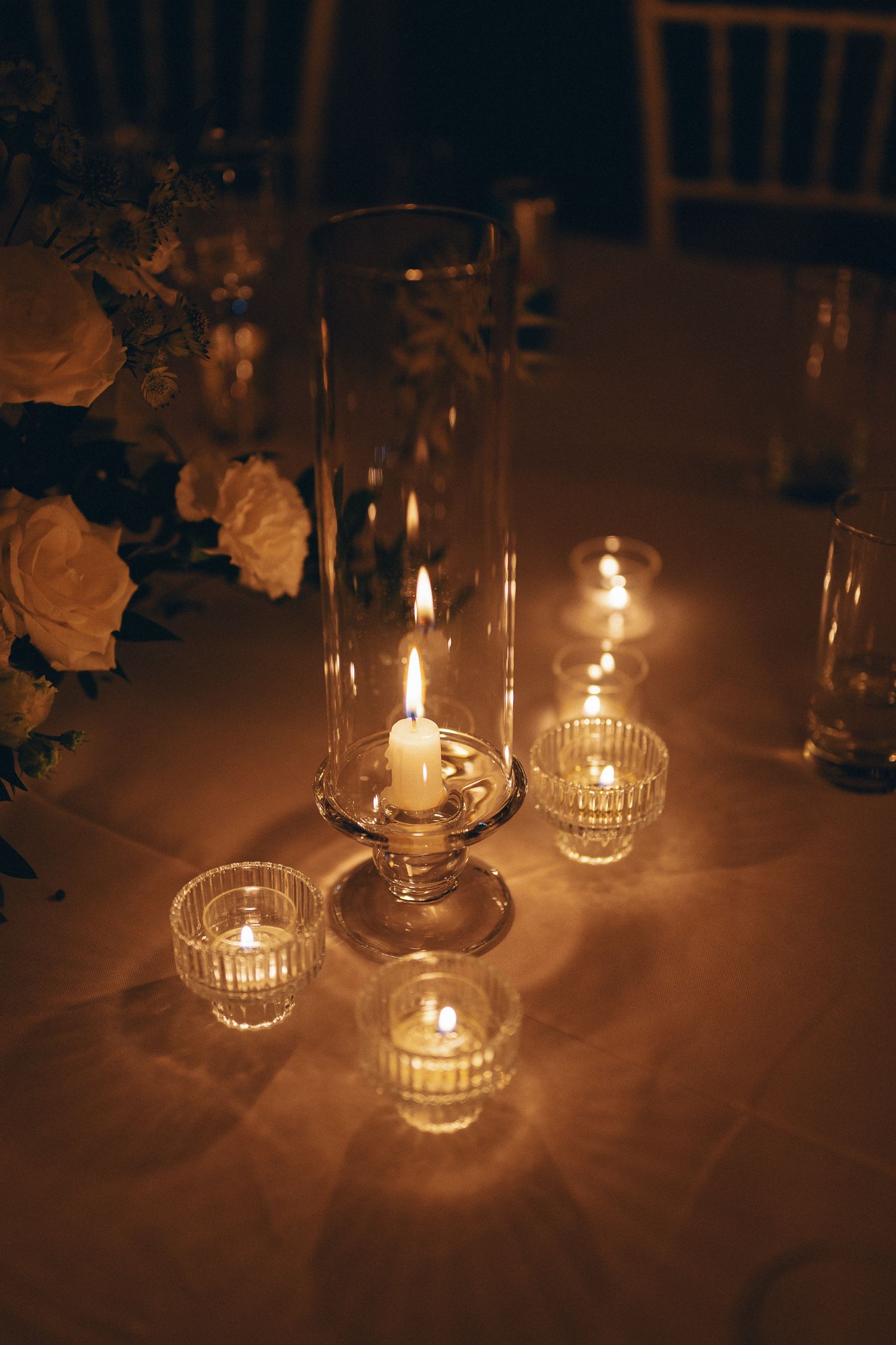 Elegant candlelit wedding table with glass hurricane candle holders and white floral arrangements in warm golden light.