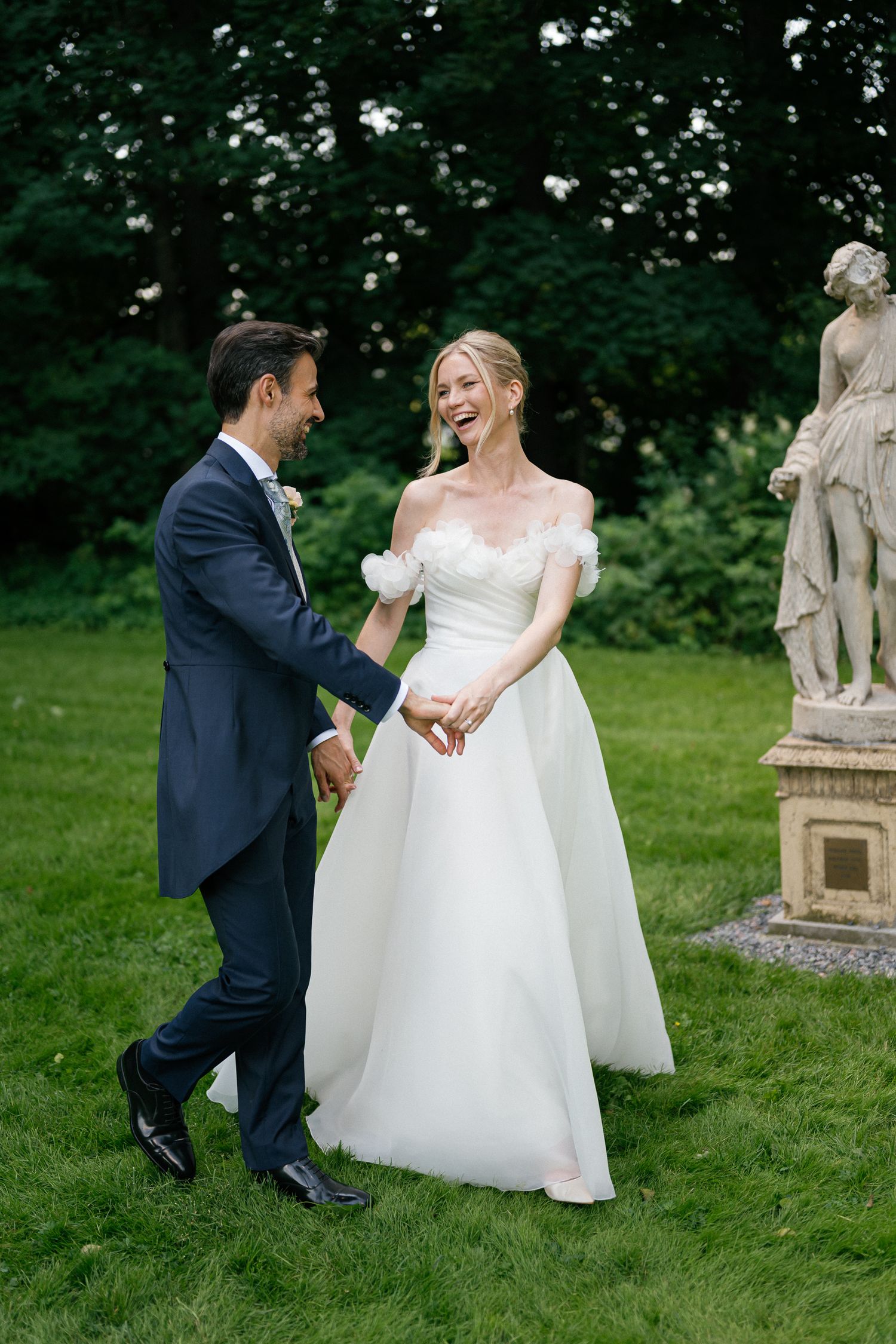 A joyful bride in a white off-shoulder gown holds hands with a groom in a navy suit on a lush green lawn.