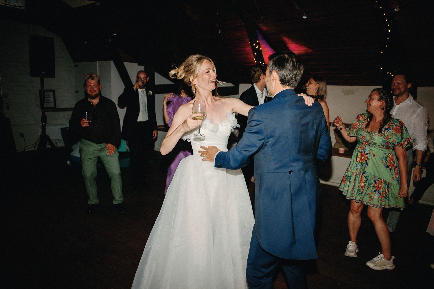 Bride in white gown dances with groom in blue suit at wedding reception while guests celebrate in background.