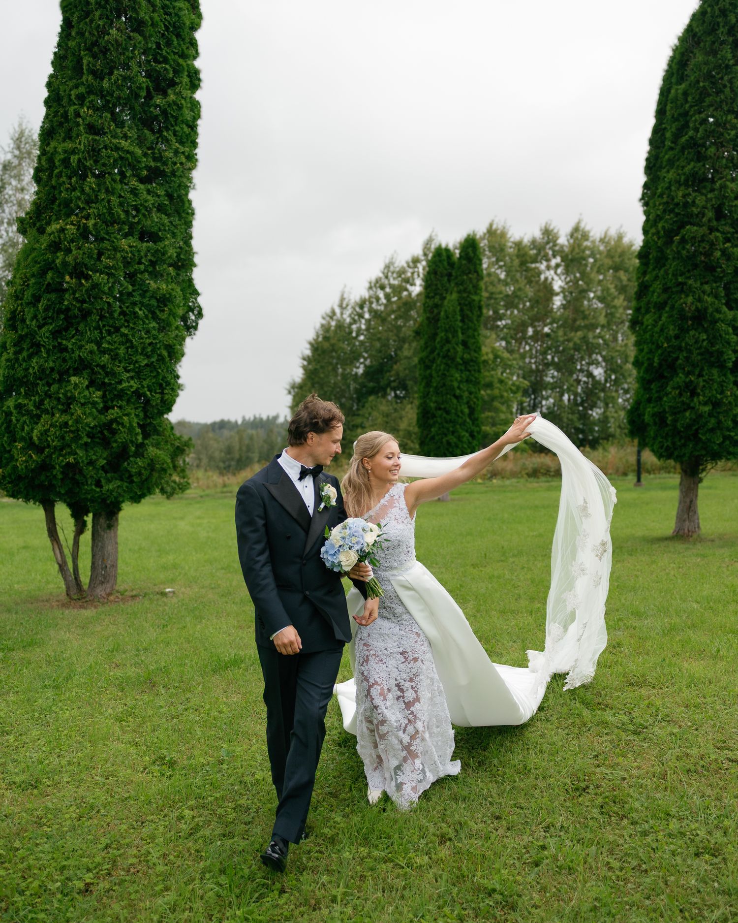Newlyweds playfully walking through a green field with tall cypress trees in the background.