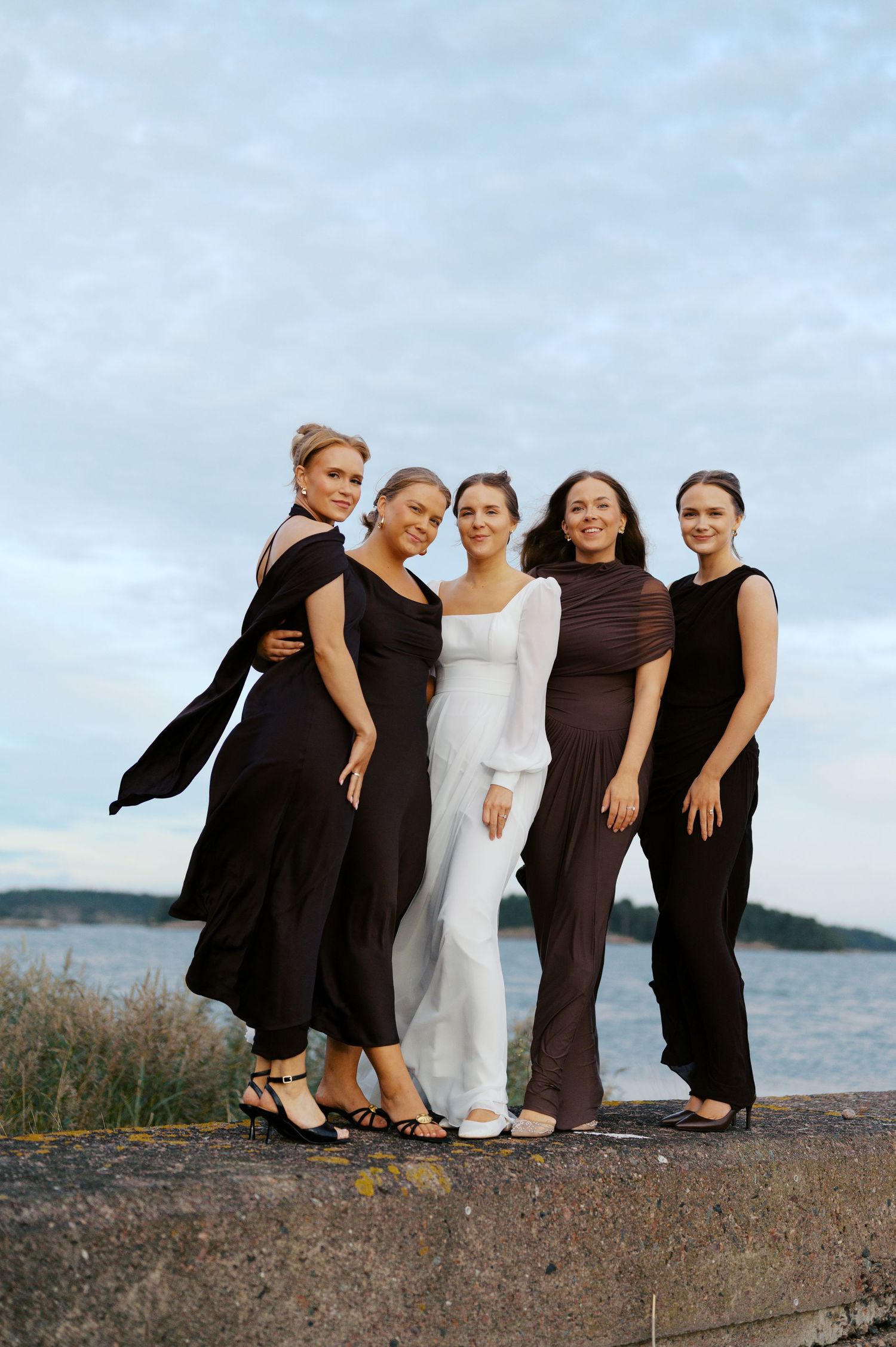 Group in black and white outfits pose together on a rocky beach at sunset with water in the background.