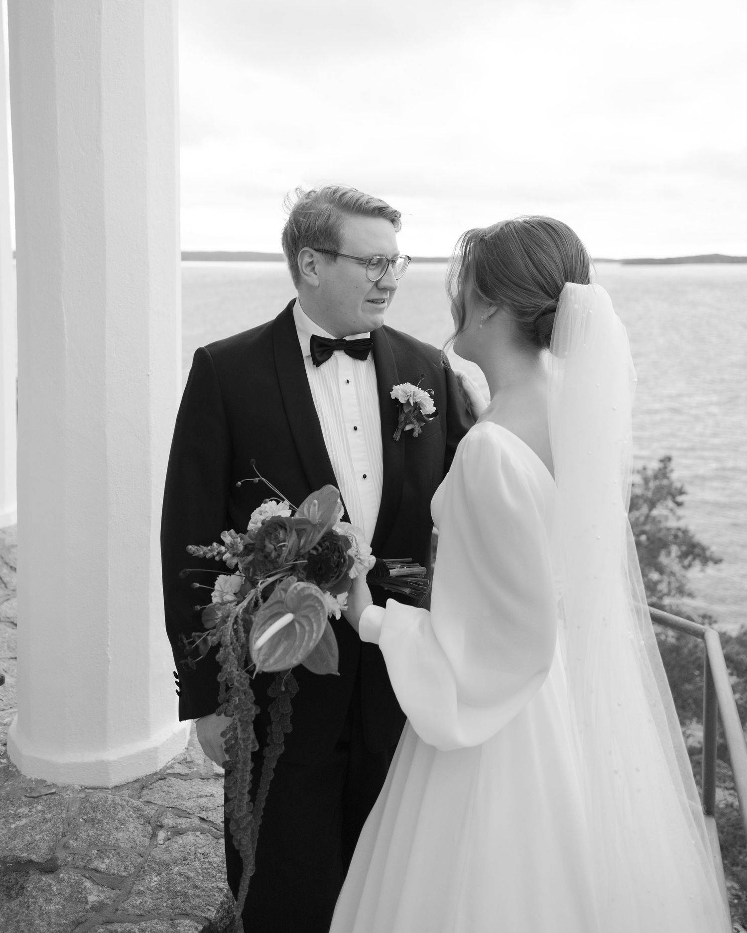 A wedding couple shares a candid moment while standing between white columns outdoors.