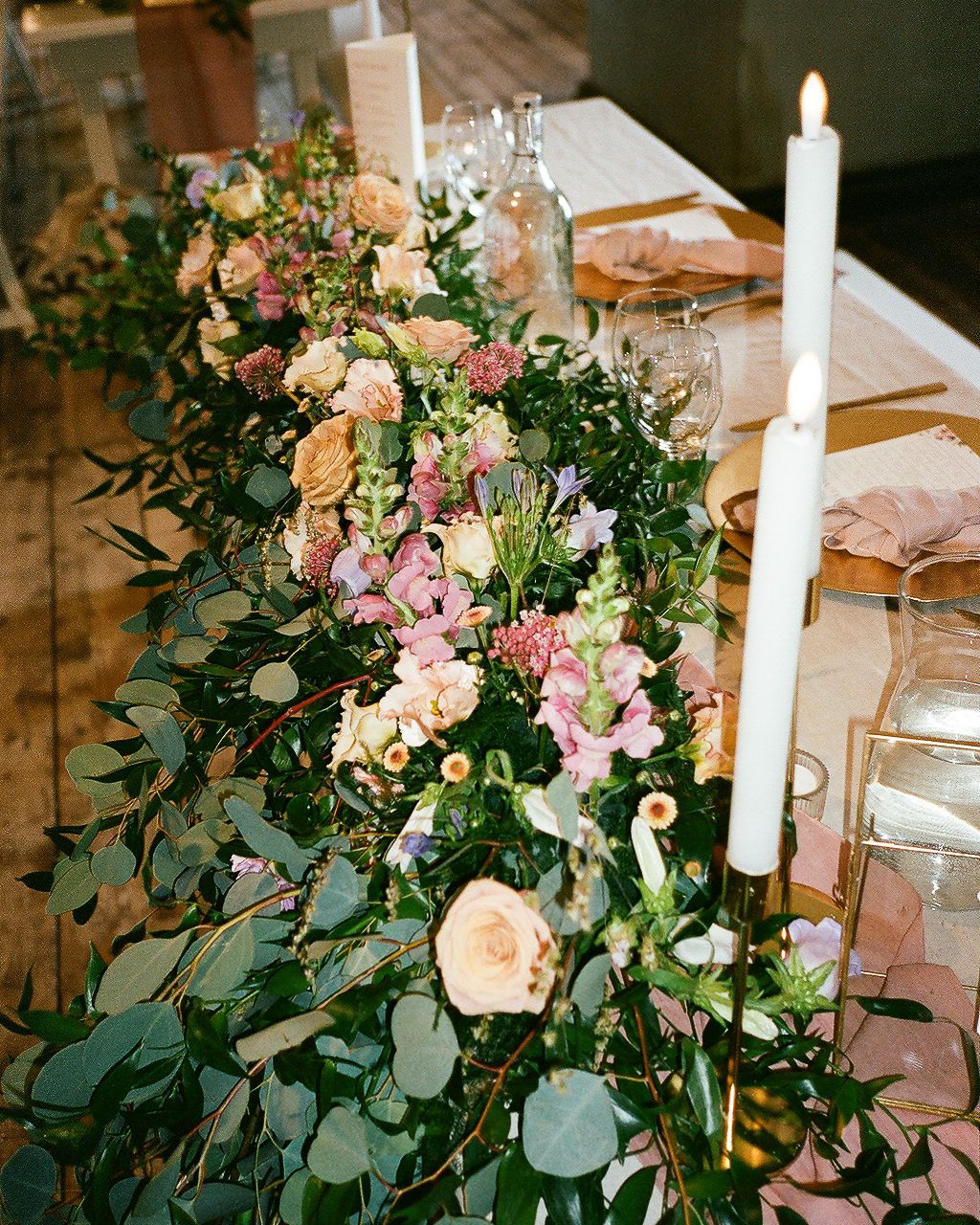 Elegant wedding table centerpiece featuring blush roses, eucalyptus greenery, and tall white taper candles in gold holders.
