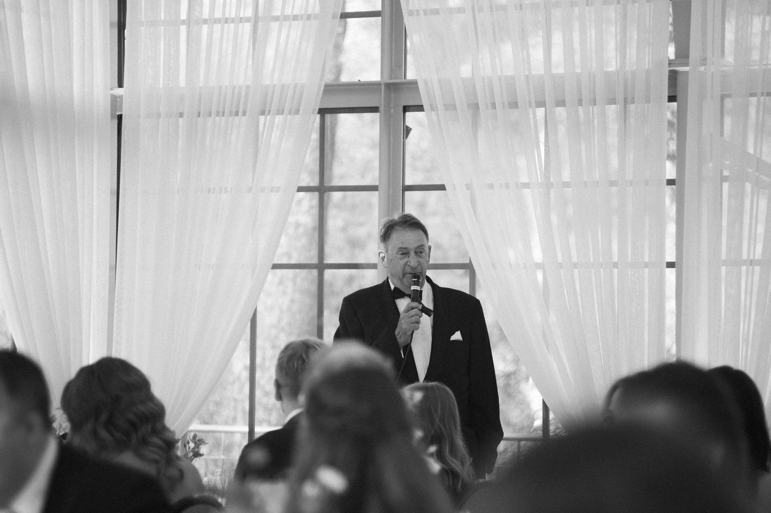 Wedding reception crowd listening to speeches in white draped venue.
