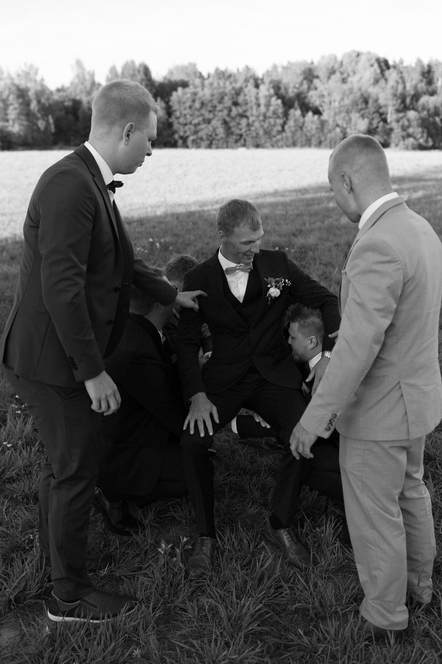 Groomsmen in formal attire stage a comedic fight sequence in a grassy field.