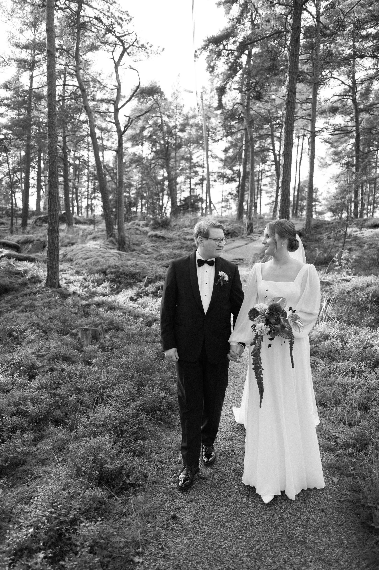 A black and white wedding photo of a couple walking together on a forest path surrounded by tall pine trees.