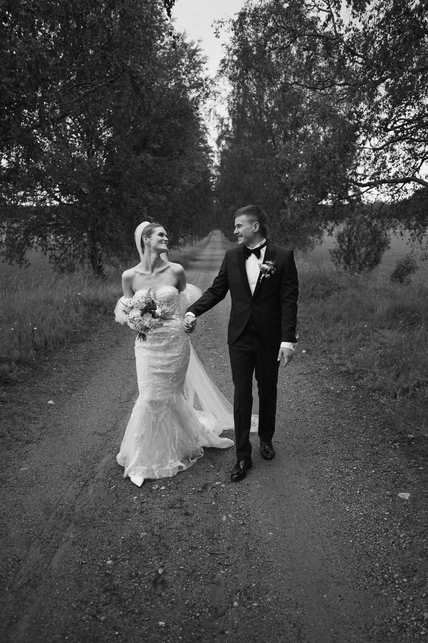 A black and white portrait of newlyweds walking together on a tree-lined path.