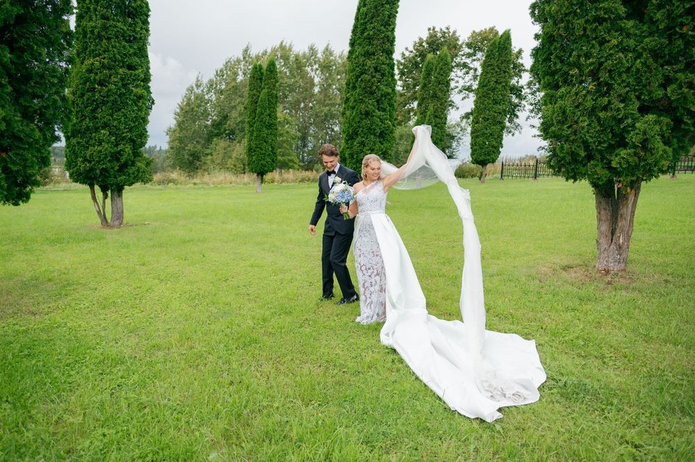 Newlyweds stroll across a lush green lawn with tall cypress trees in the background.