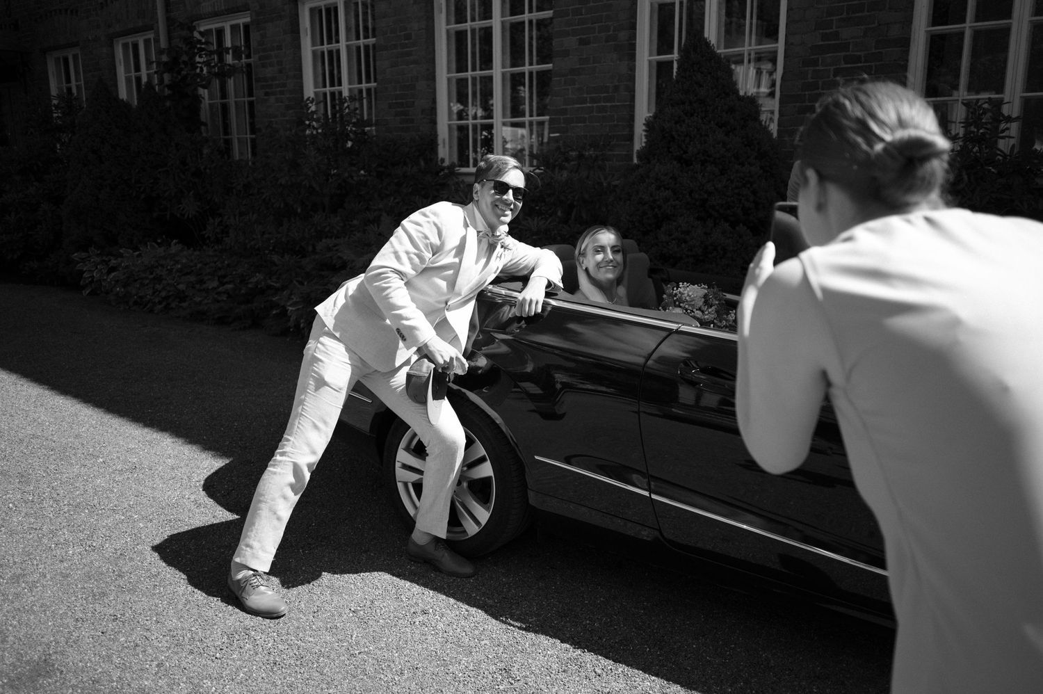 Black and white image of wedding scene with car and brick building backdrop.