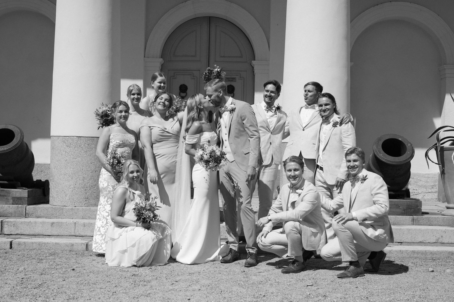 Wedding party members sit casually on steps of elegant columned building in light formal attire.