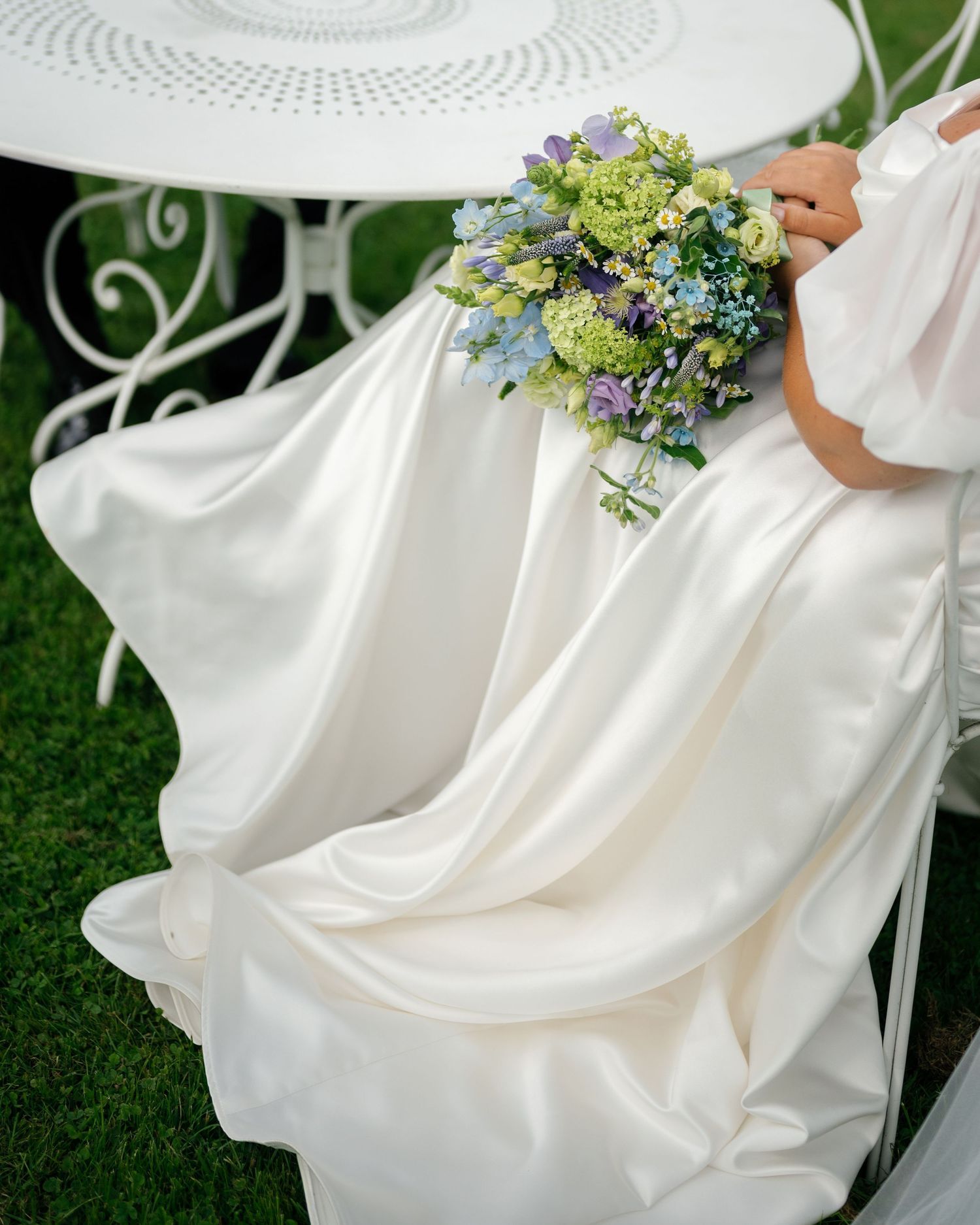 Close-up of flowing white wedding dress with delicate blue and green floral bouquet.