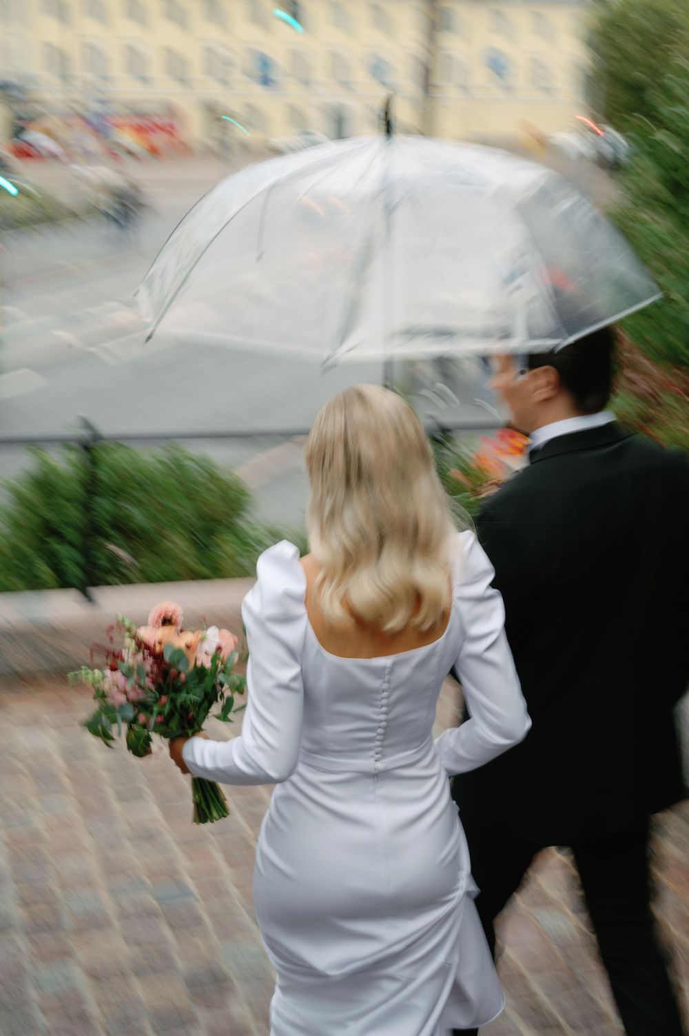 Wedding couple walks under clear umbrella on rainy day with floral bouquet.