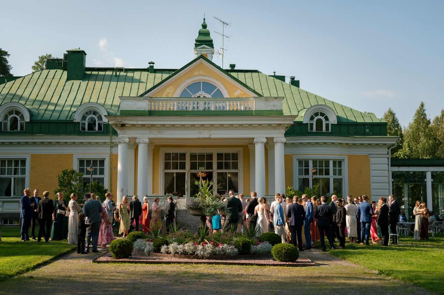 A panoramic view of a historic mansion with green roof and white columns surrounded by manicured gardens and visitors.