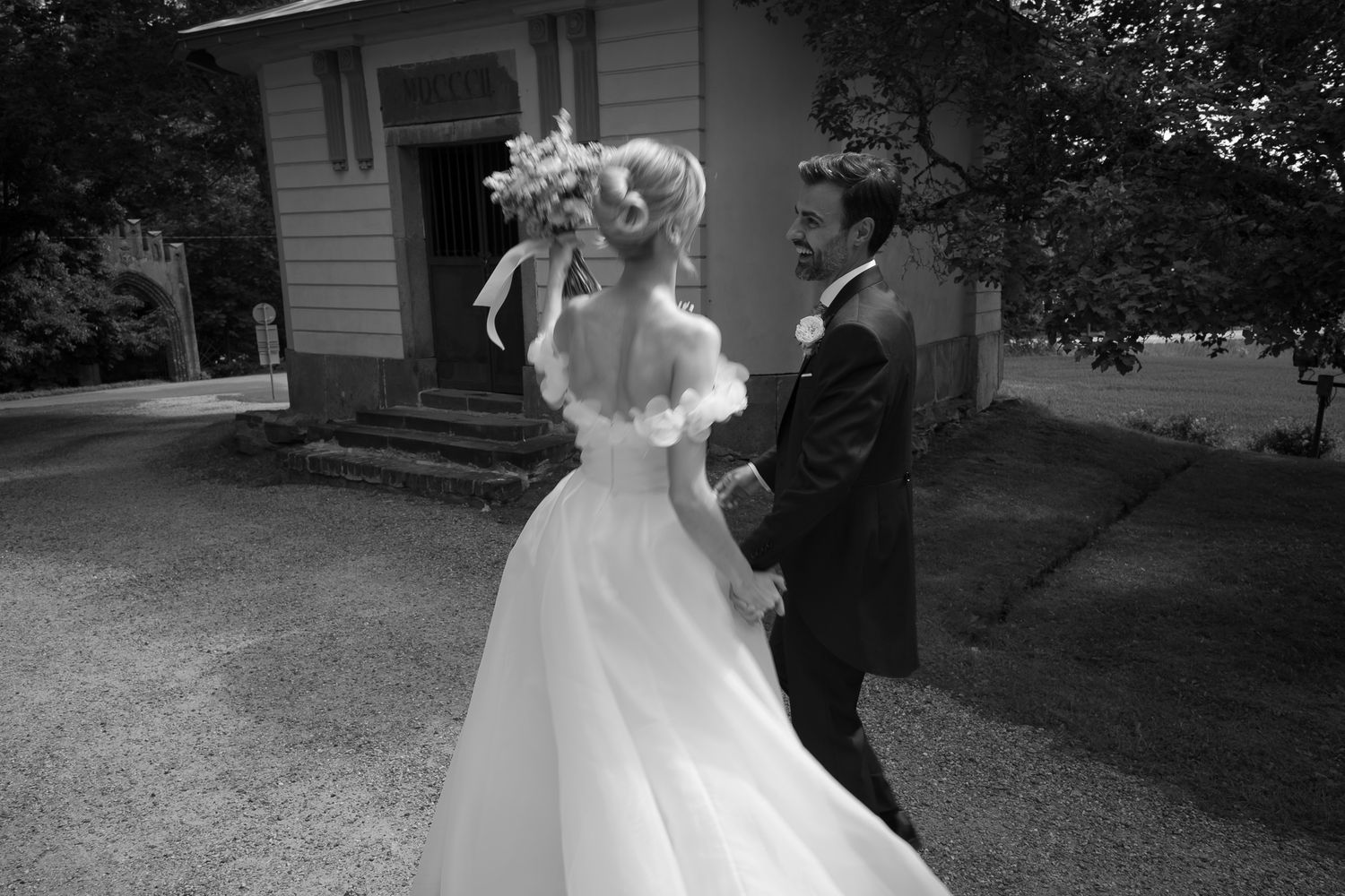 Black and white photo of a bride in an off-shoulder gown holding a bouquet, walking with a groom in a suit outdoors.