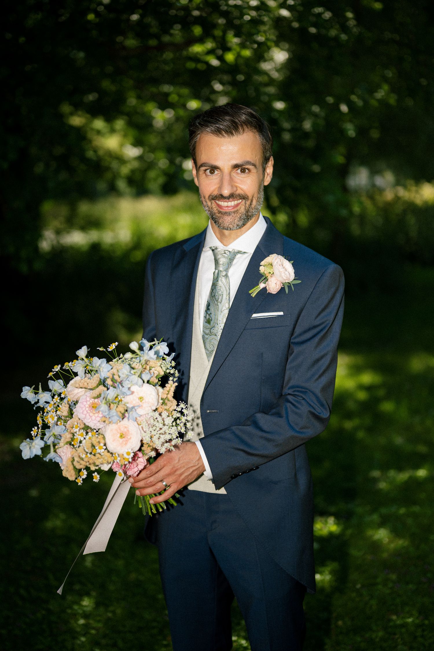 Smiling groom in navy suit holding bridal bouquet of pastel flowers with boutonniere outdoors.