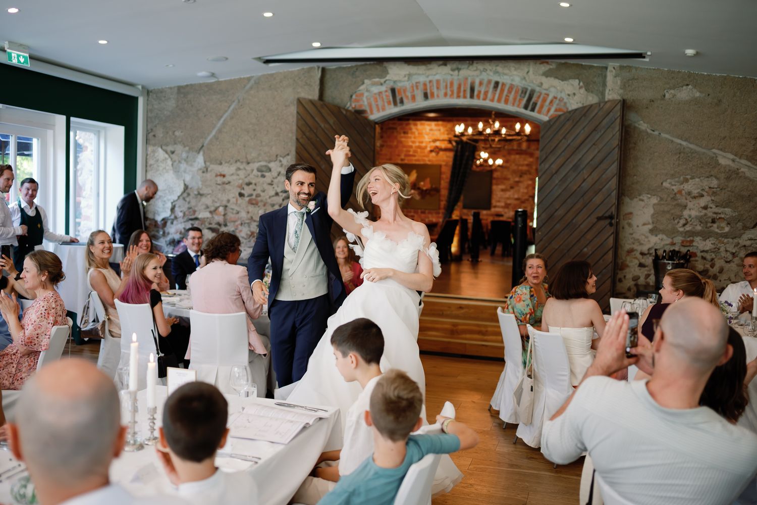 Joyful bride and groom celebrate their grand reception entrance as guests applaud in a rustic stone venue.