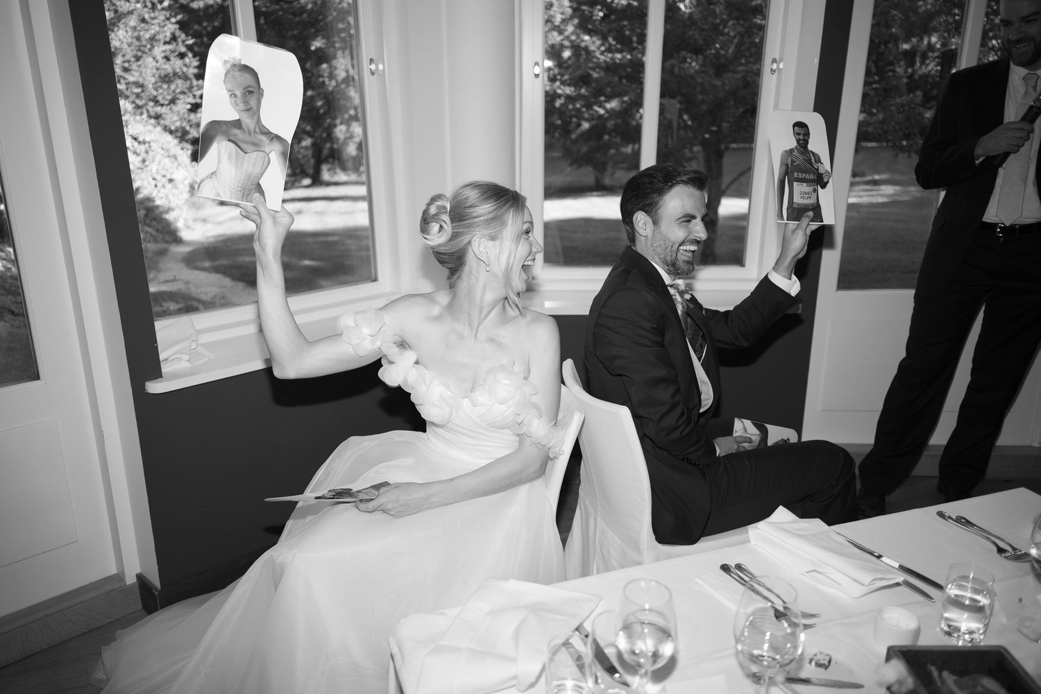 Black and white photo of a bride and groom holding up pictures at their wedding reception table.