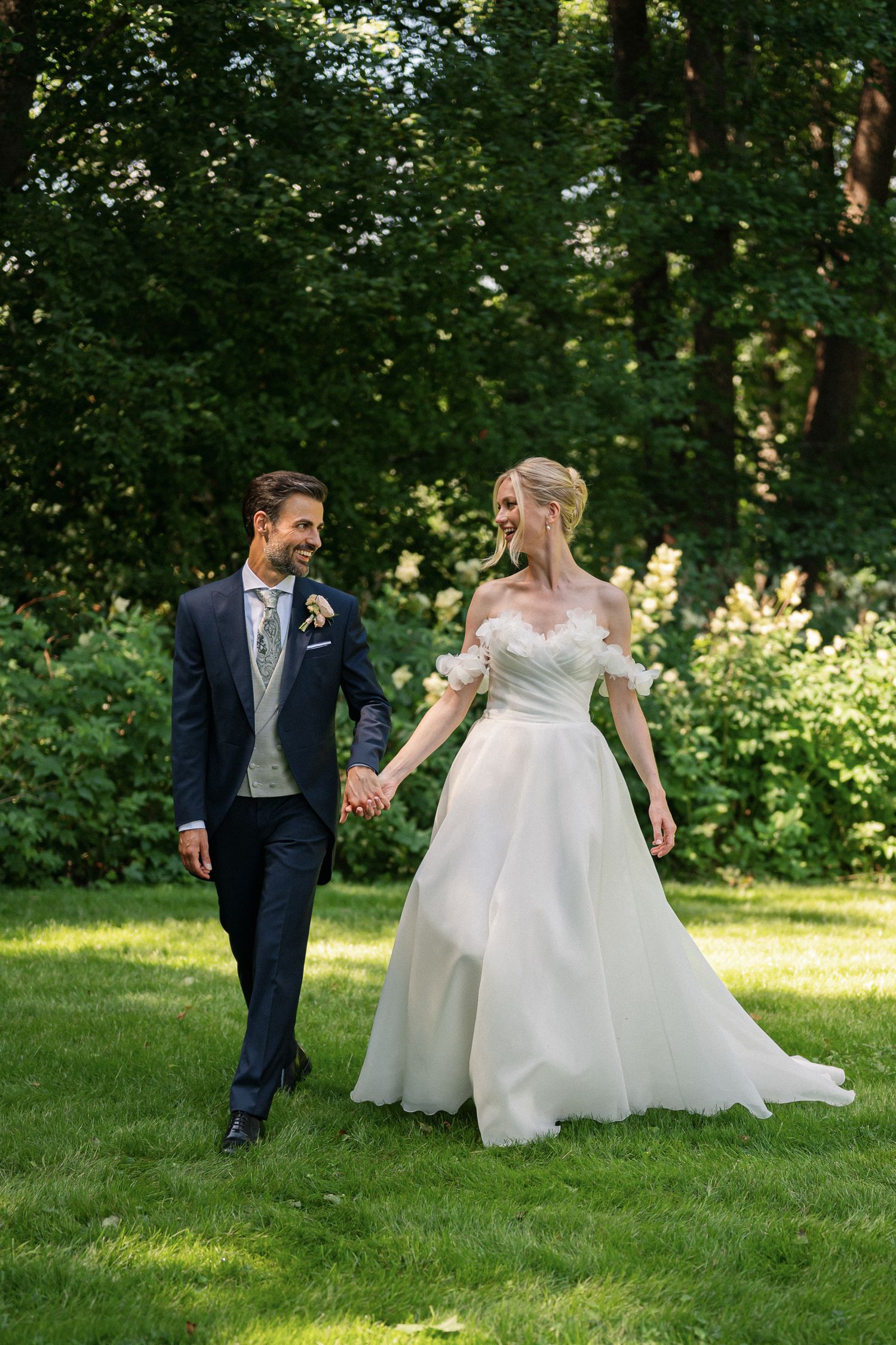 A bride and groom walk hand-in-hand through a lush green garden, smiling at each other on their wedding day.