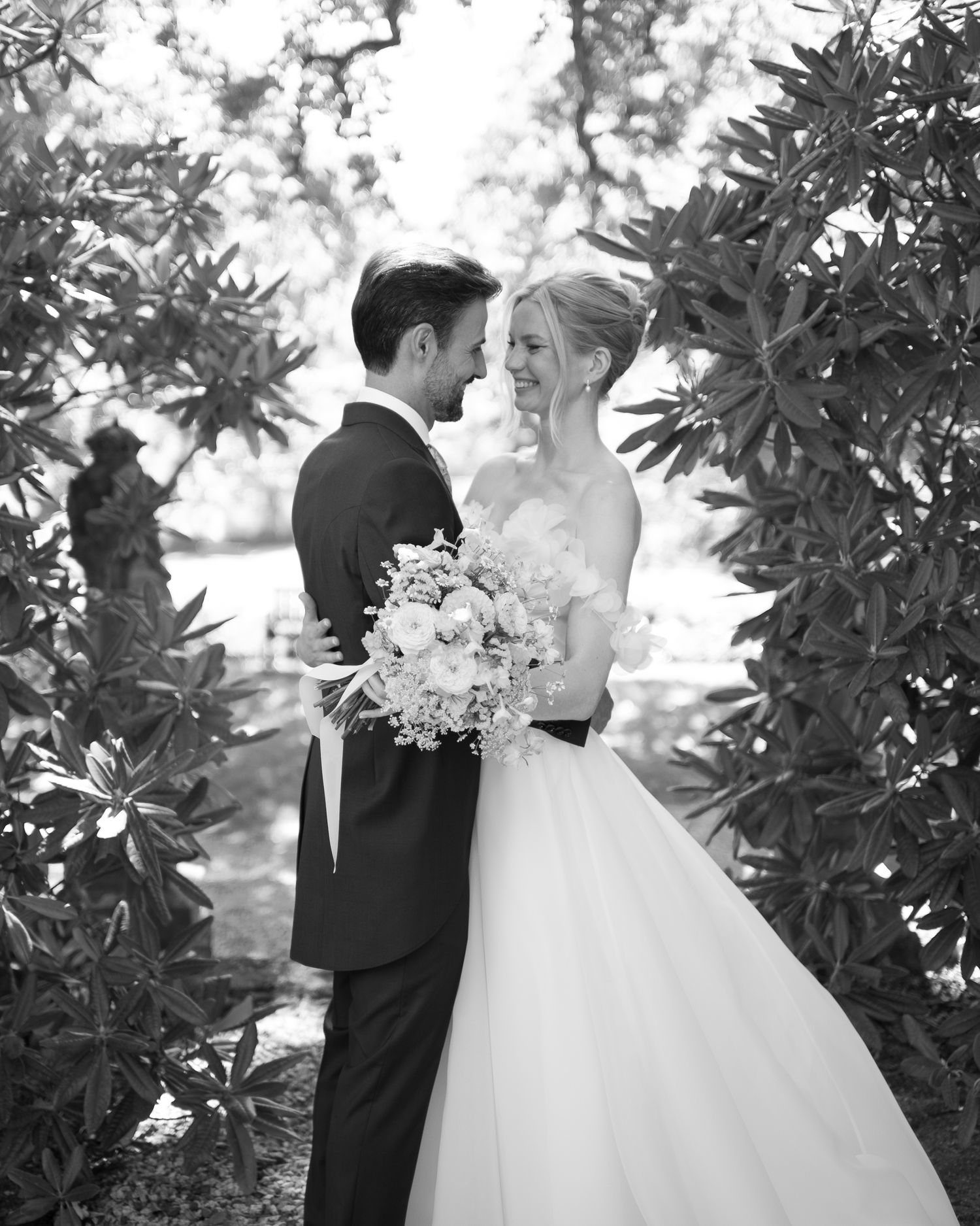 Black and white wedding photo of smiling bride holding bouquet and groom in tuxedo embracing among lush garden foliage.