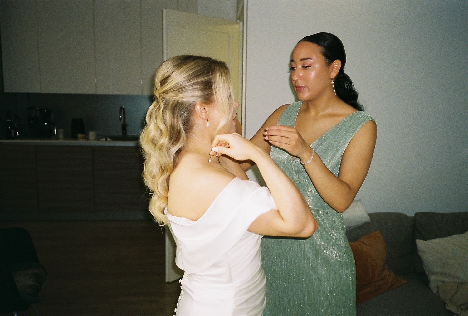 Two women in elegant dresses, one blonde in white, the other in green, adjusting jewelry indoors.