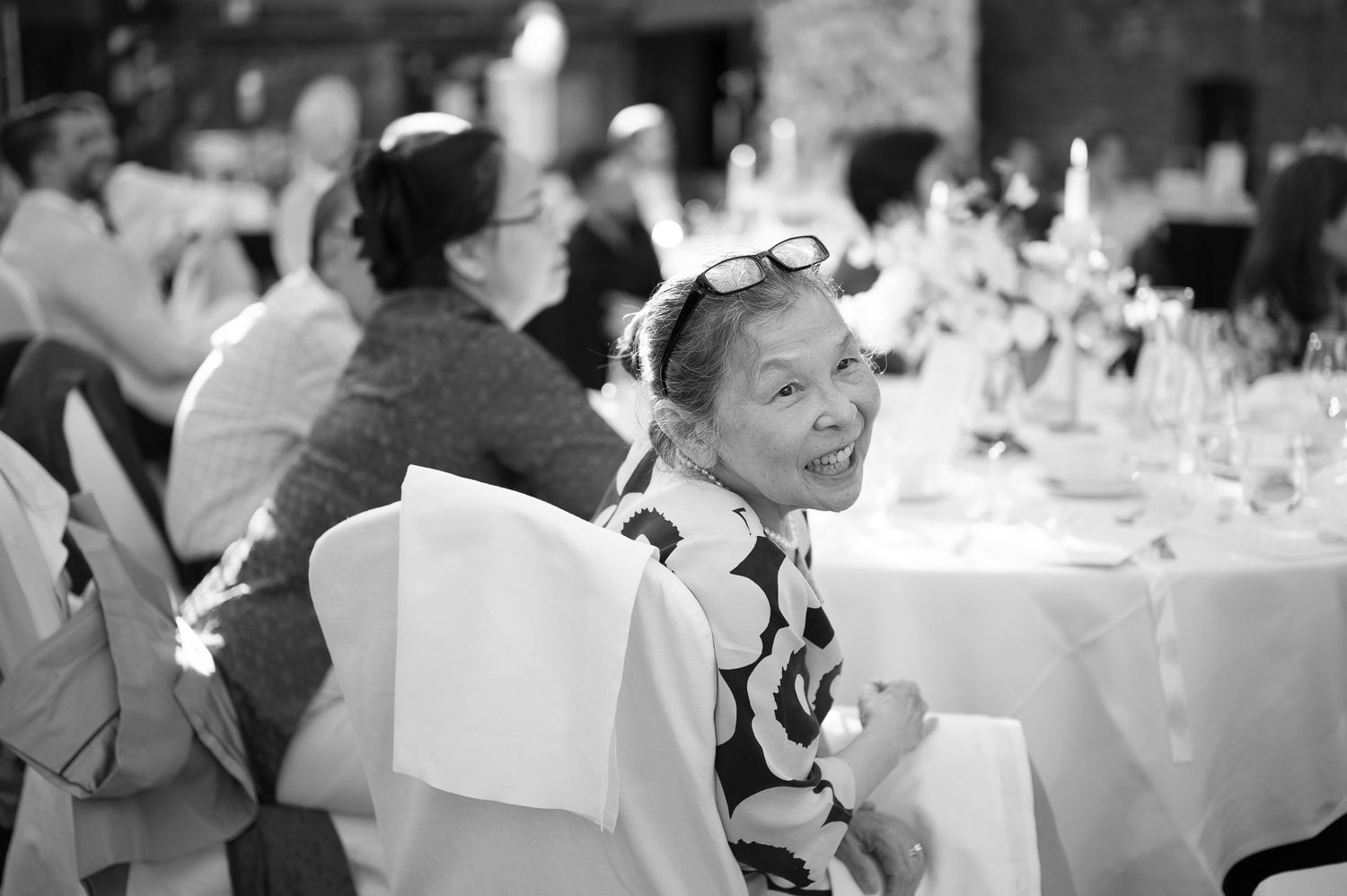 Woman smiling at a formal dinner event, wearing a patterned dress with sunglasses on her head, black and white photo.