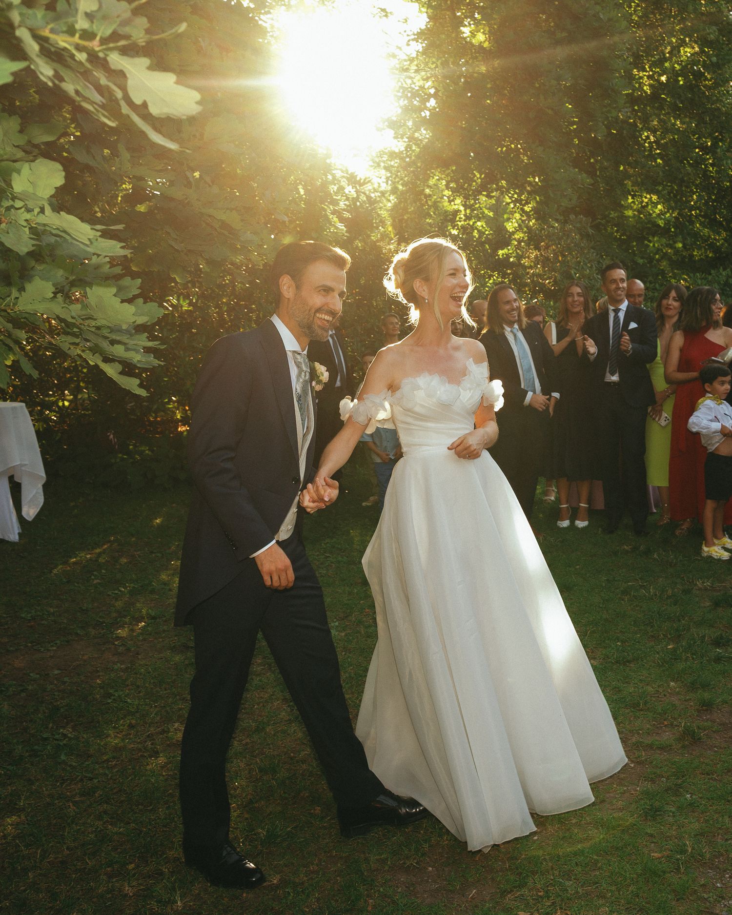 A bride and groom walk hand in hand outdoors at golden hour, surrounded by cheering guests in a lush garden setting.