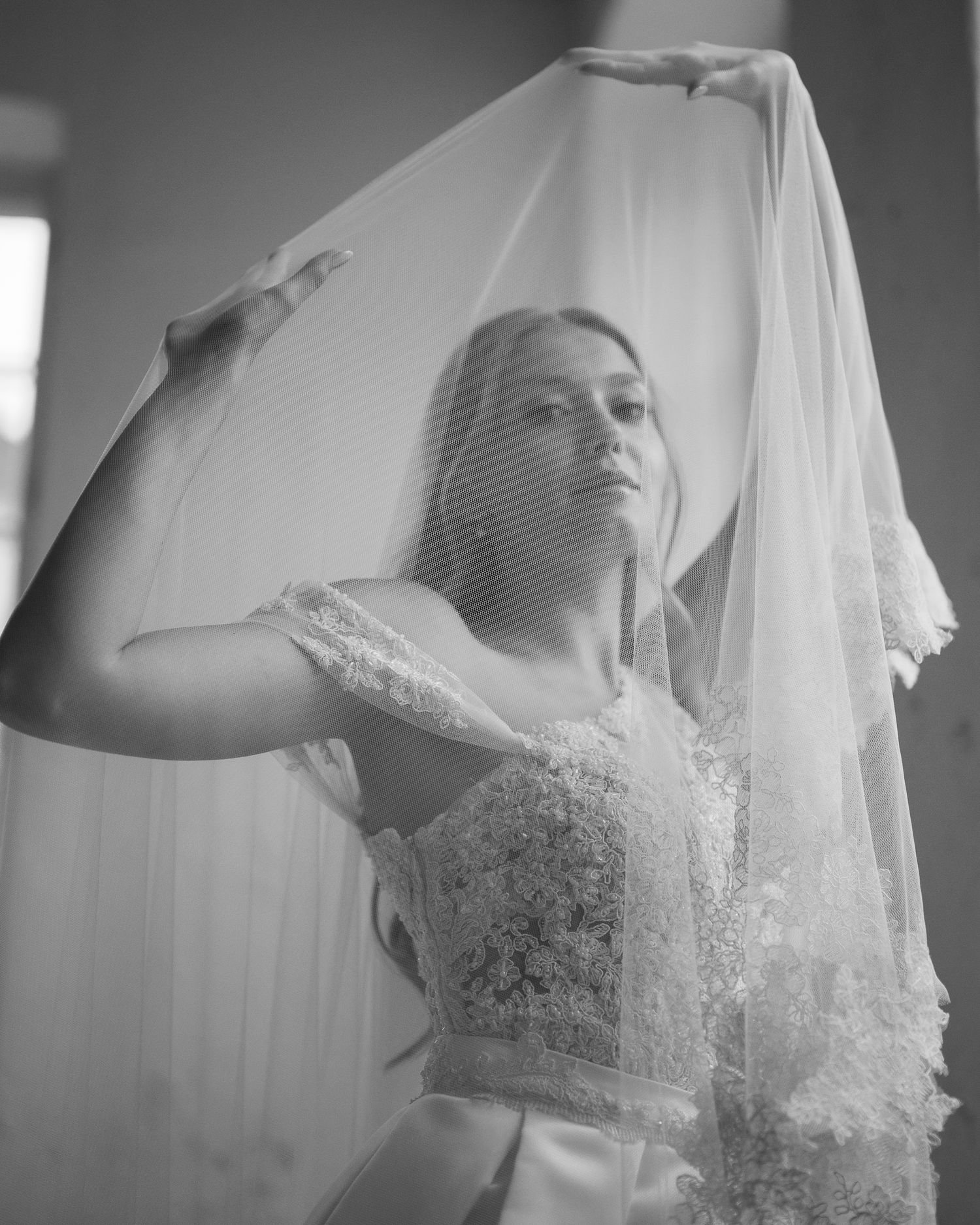Black and white photo of a bride in lace wedding dress lifting her veil gracefully indoors.