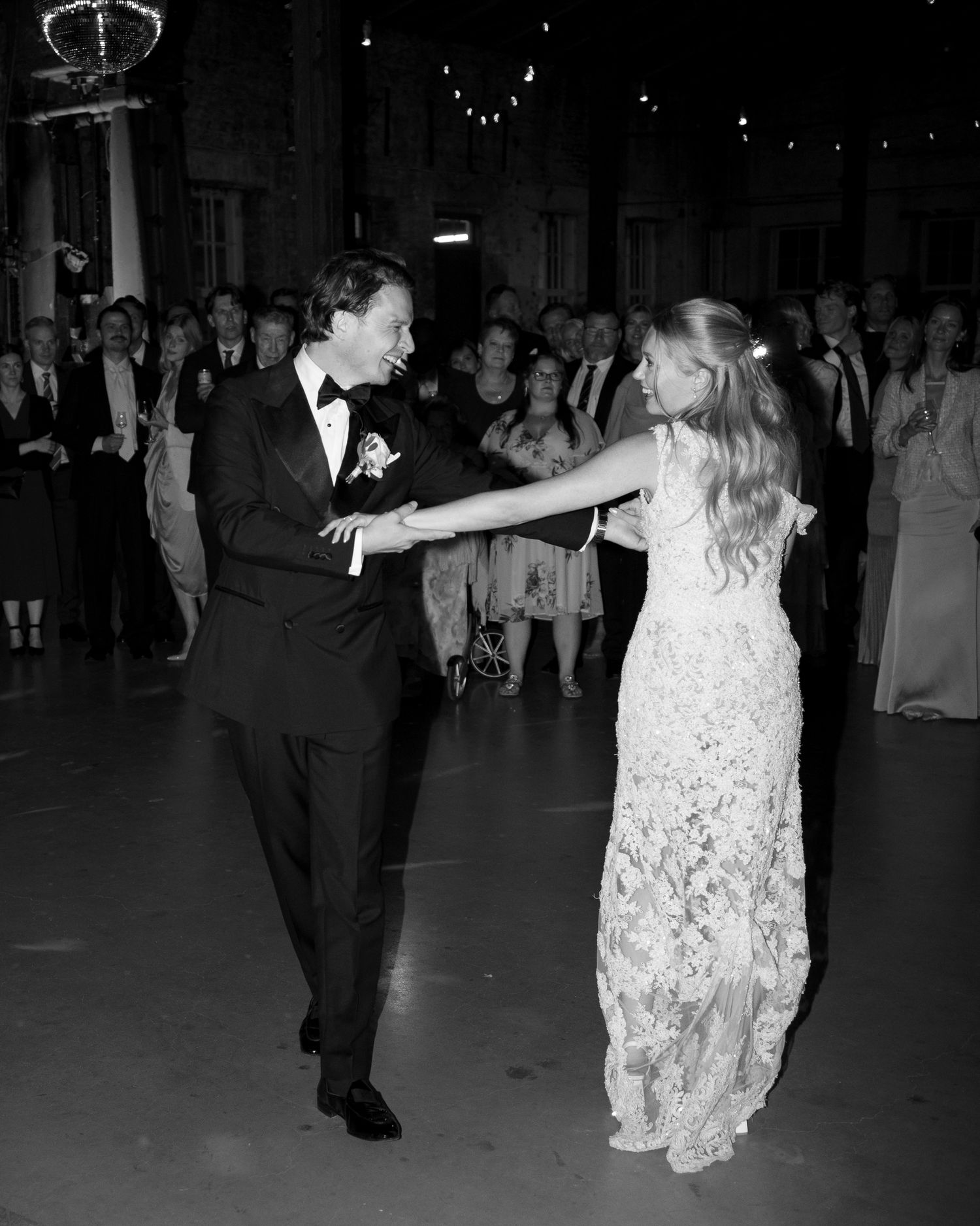 A couple dances together at a formal event, with the woman wearing a white lace dress and the man in a black tuxedo.