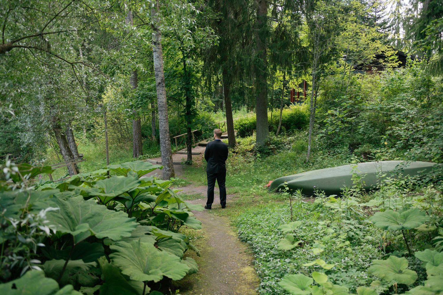 A figure stands on a wooded path surrounded by lush greenery and tall trees in a tranquil forest setting.