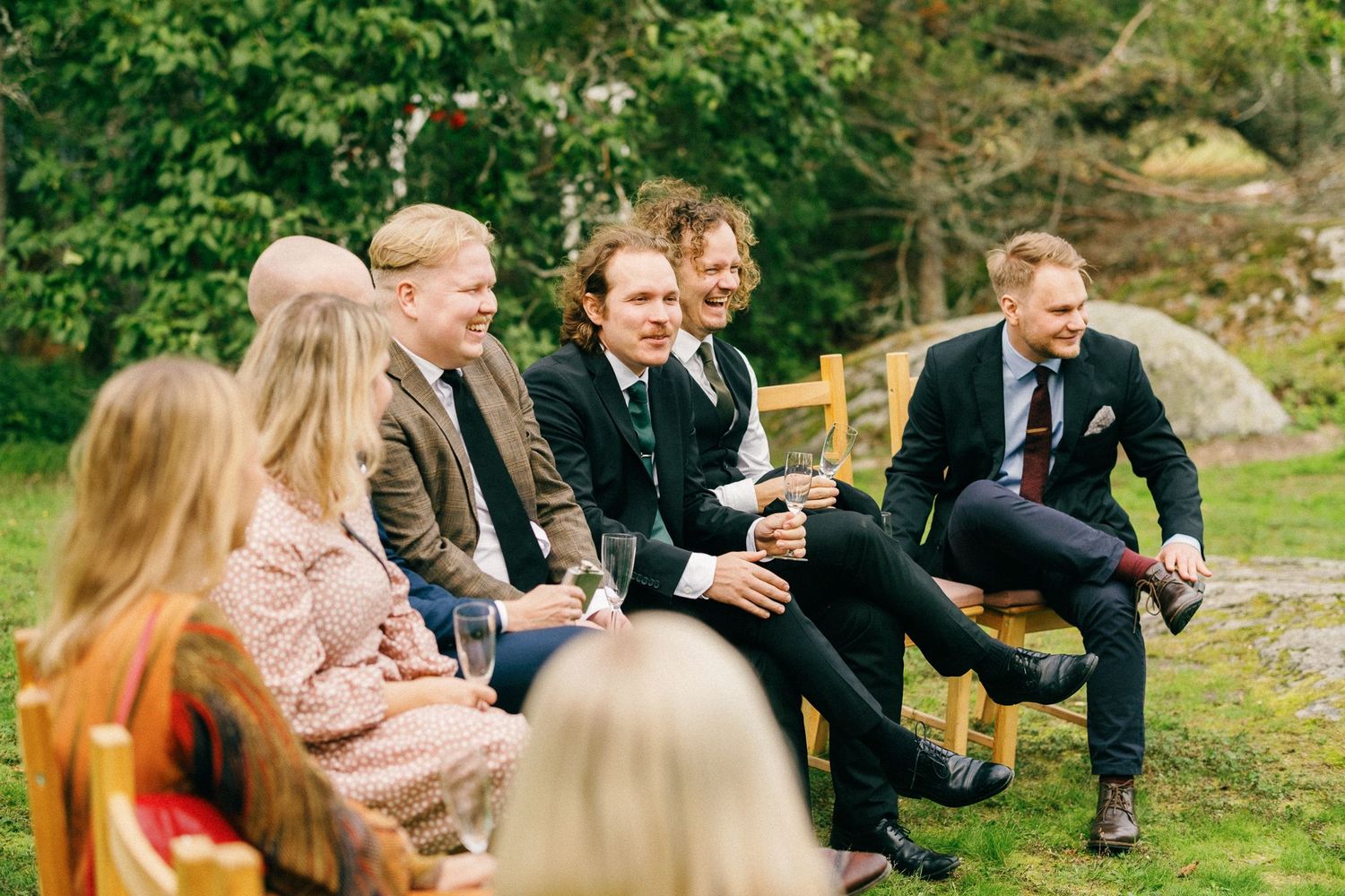 A group of guests sharing laughs while seated outdoors on wooden chairs during a casual gathering.