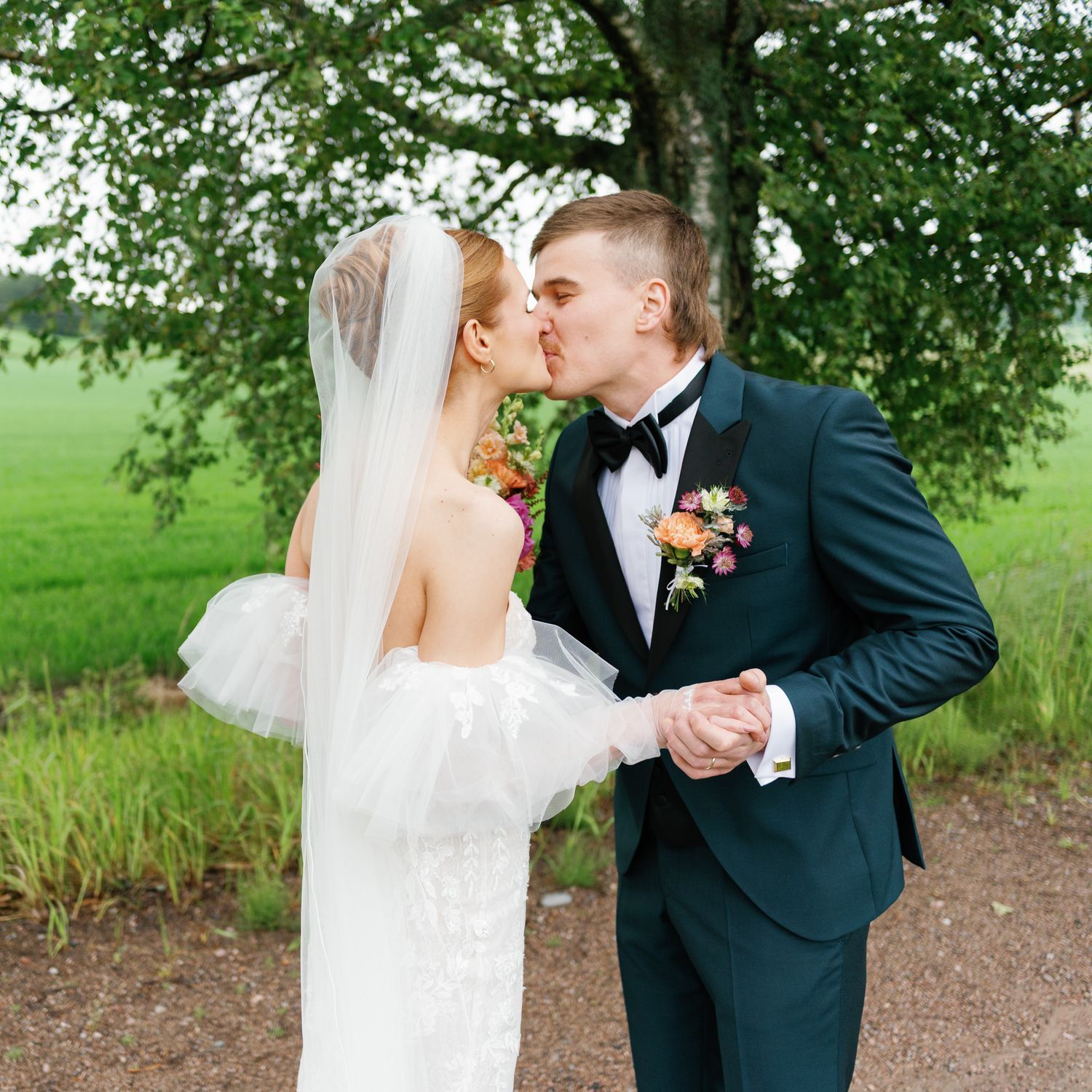 A tender wedding kiss captured under a leafy tree with rolling green fields in the background.