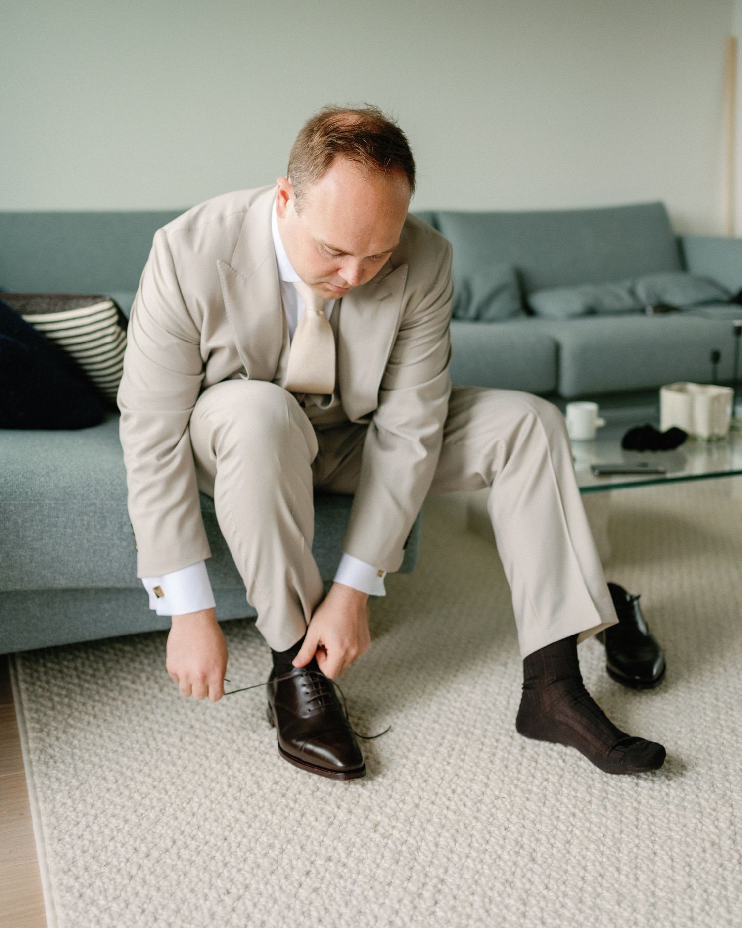 A groom in a beige suit sits on a carpet tying his brown dress shoes before his wedding ceremony.