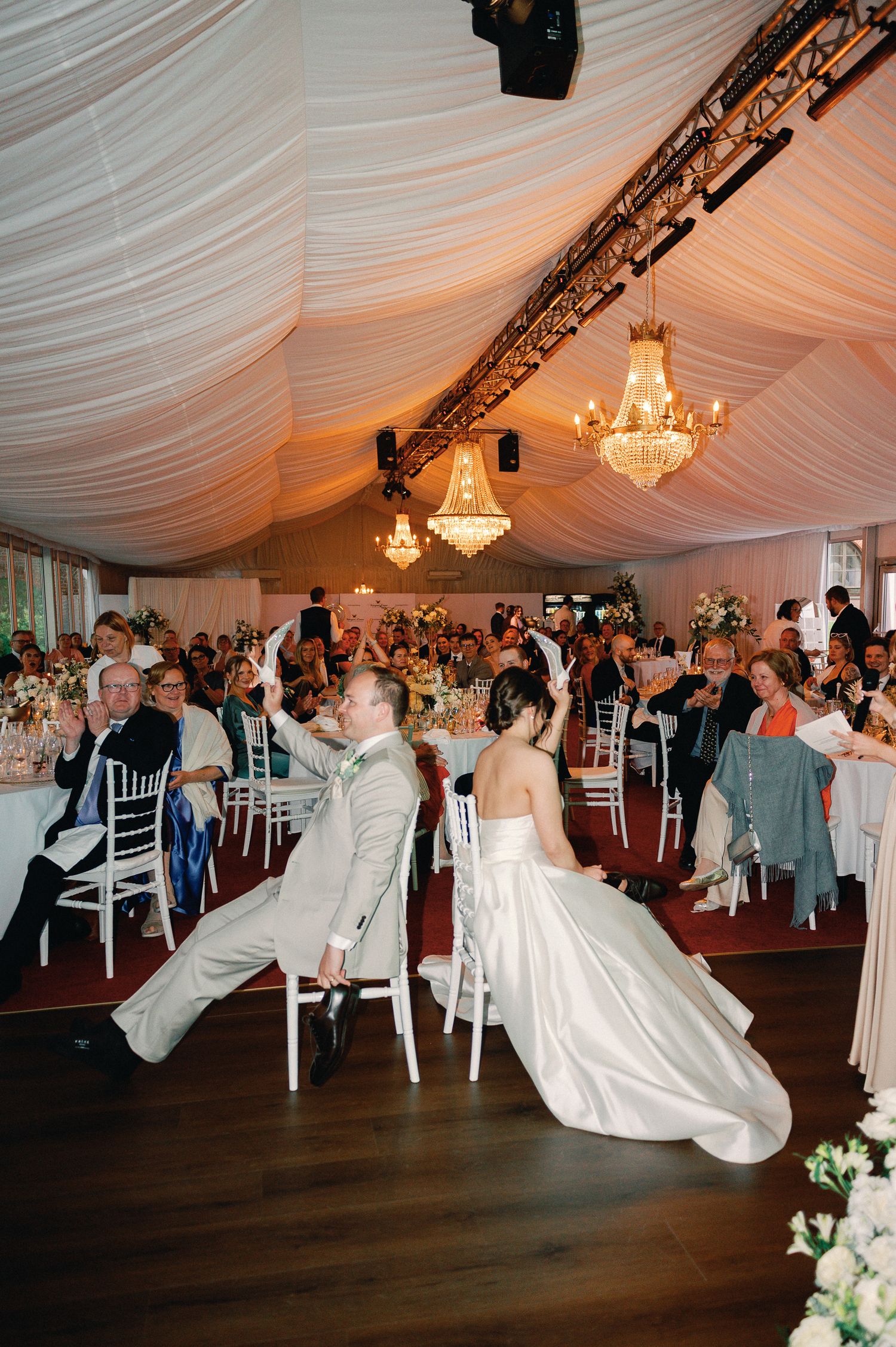 Bride and groom lean back in chairs during wedding reception in elegant tent venue with chandeliers and guests.