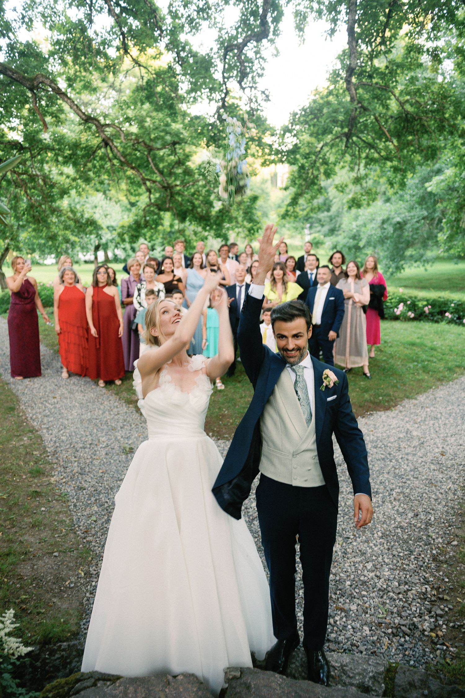 Joyful bride and groom raising hands on gravel path with wedding guests celebrating in lush green garden.