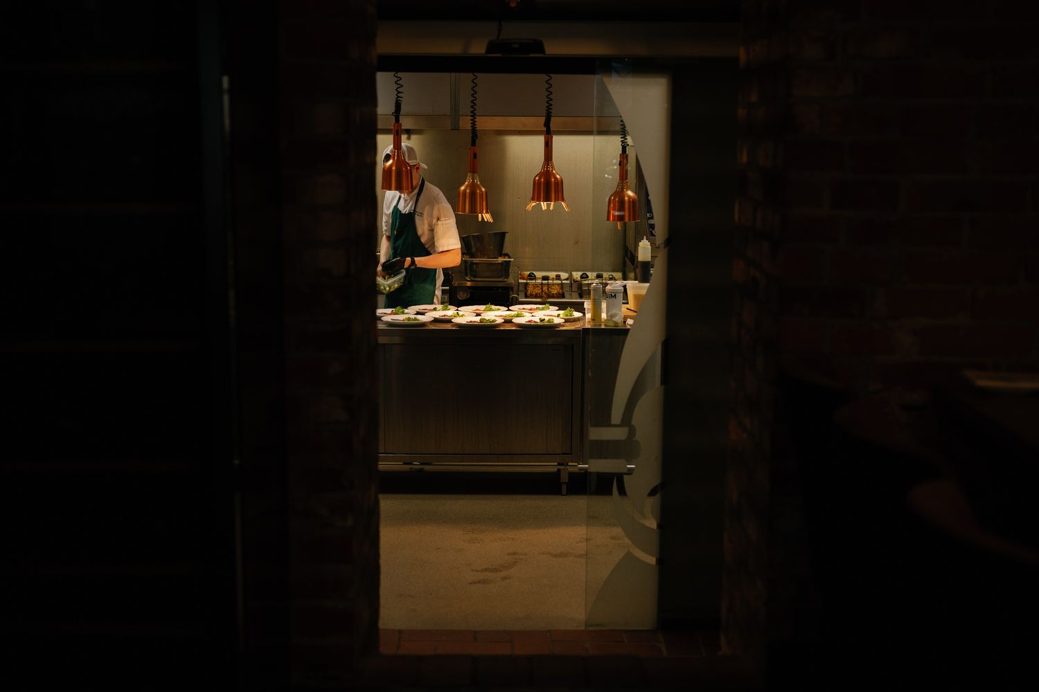 A chef works in a warmly lit restaurant kitchen, visible through a dark doorway with hanging copper lights.