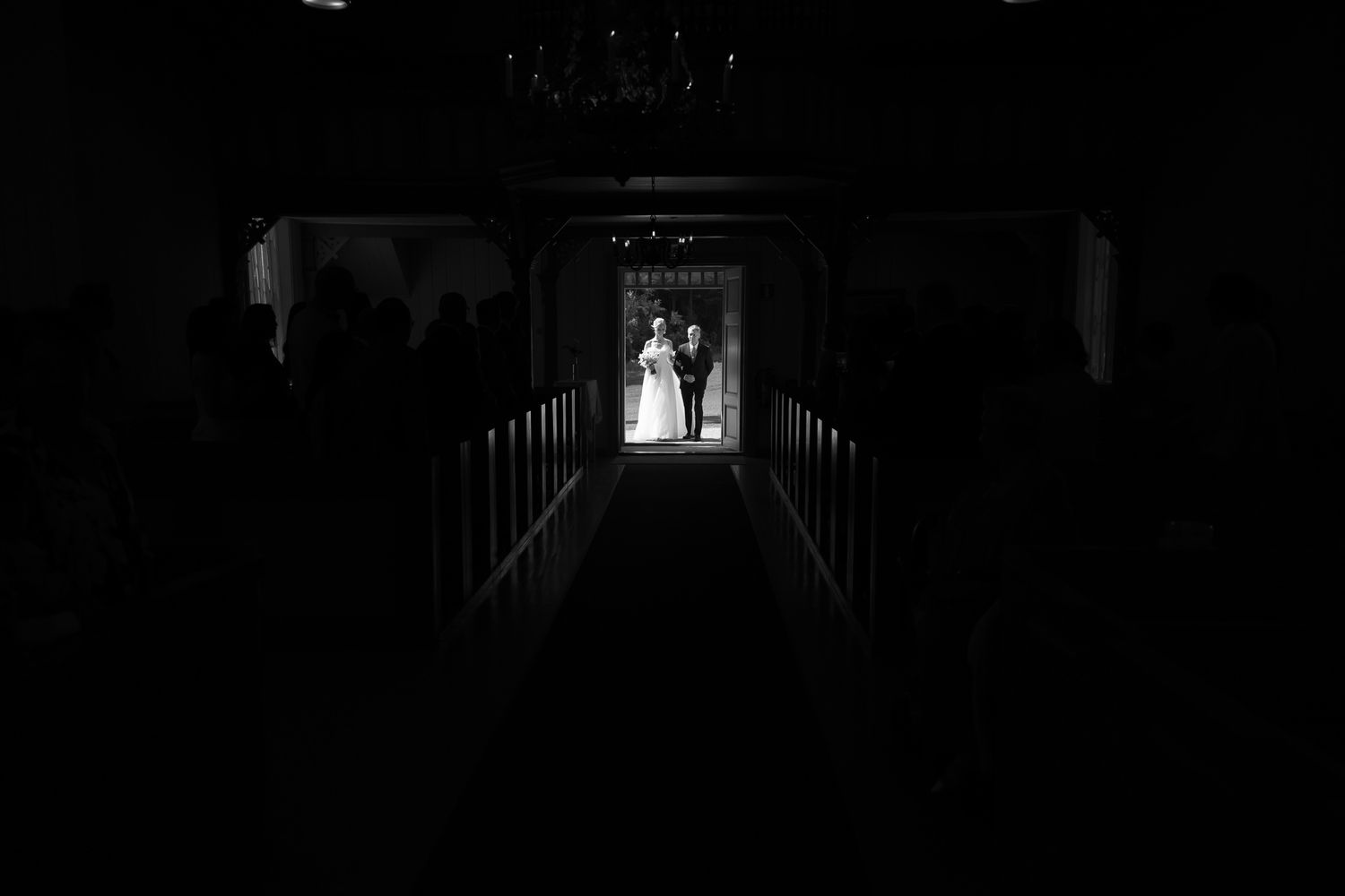 Black and white photo of a wedding couple framed in a bright doorway at the end of a dark church aisle.
