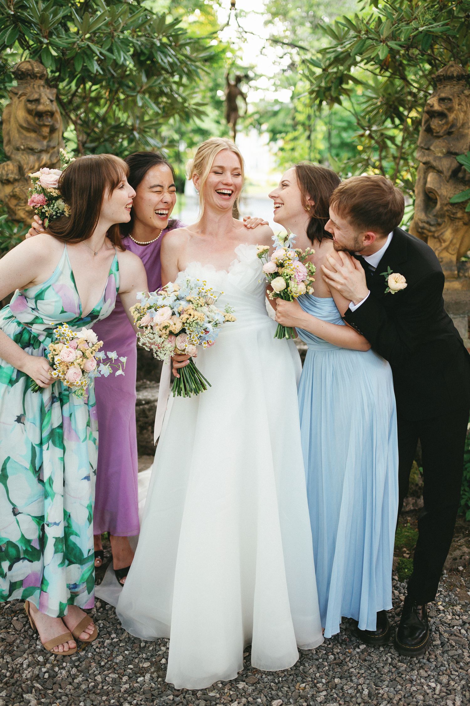 A laughing bride in white holds a bouquet surrounded by bridesmaids in colorful dresses and a groomsman in a garden.