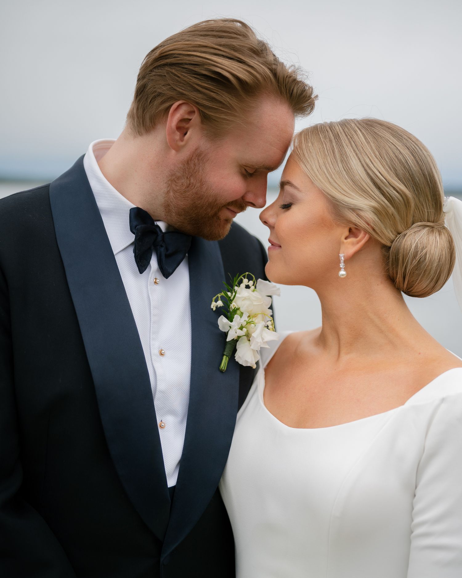 A bride and groom in formal wedding attire share an intimate moment, touching foreheads outdoors near water.