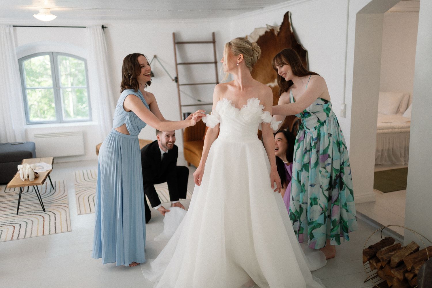 Bridesmaids in blue and floral dresses help a bride in a white gown prepare in a bright, modern wedding venue.