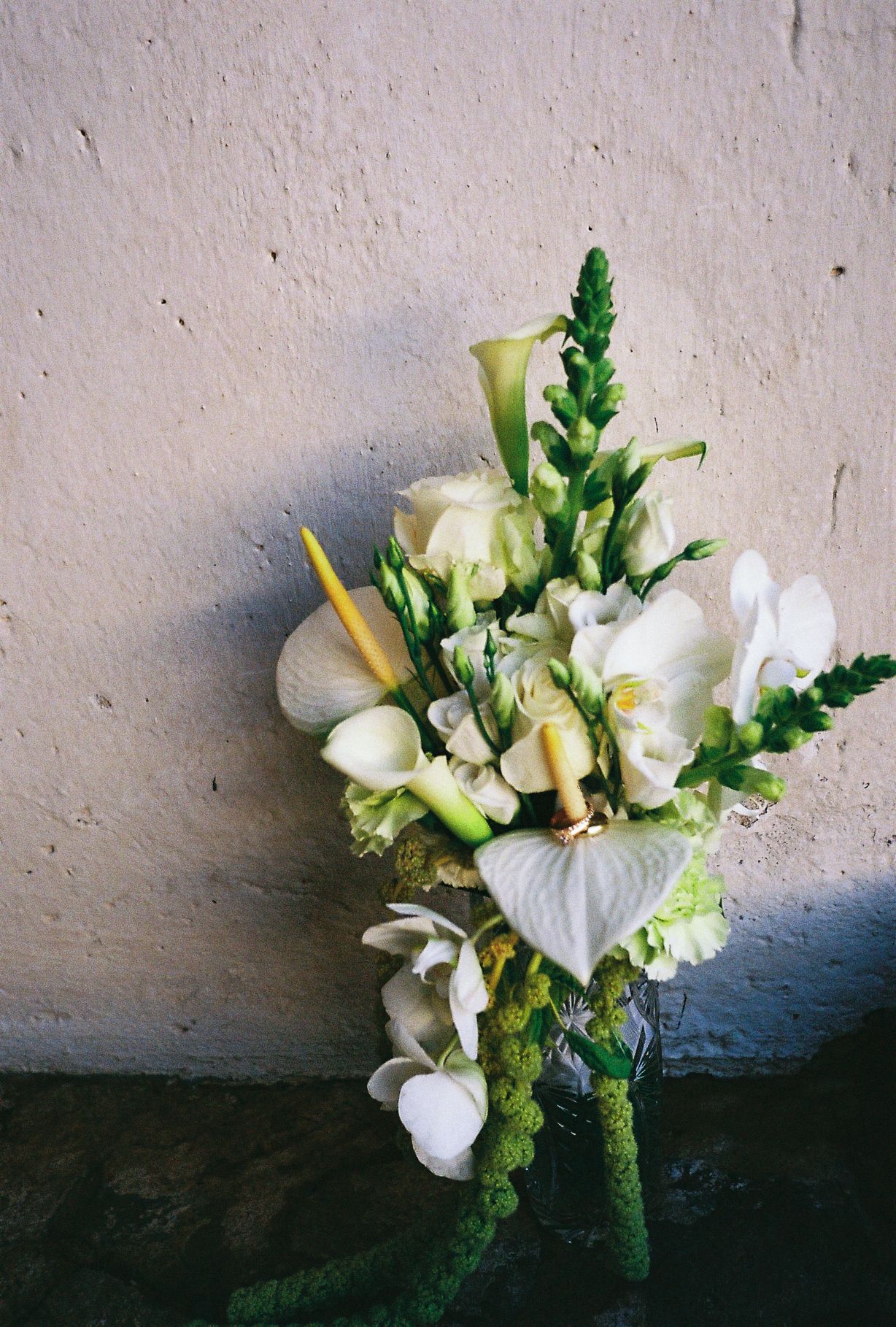 White floral boutonniere with calla lilies, orchids, and green accents leaning against a textured gray wall.