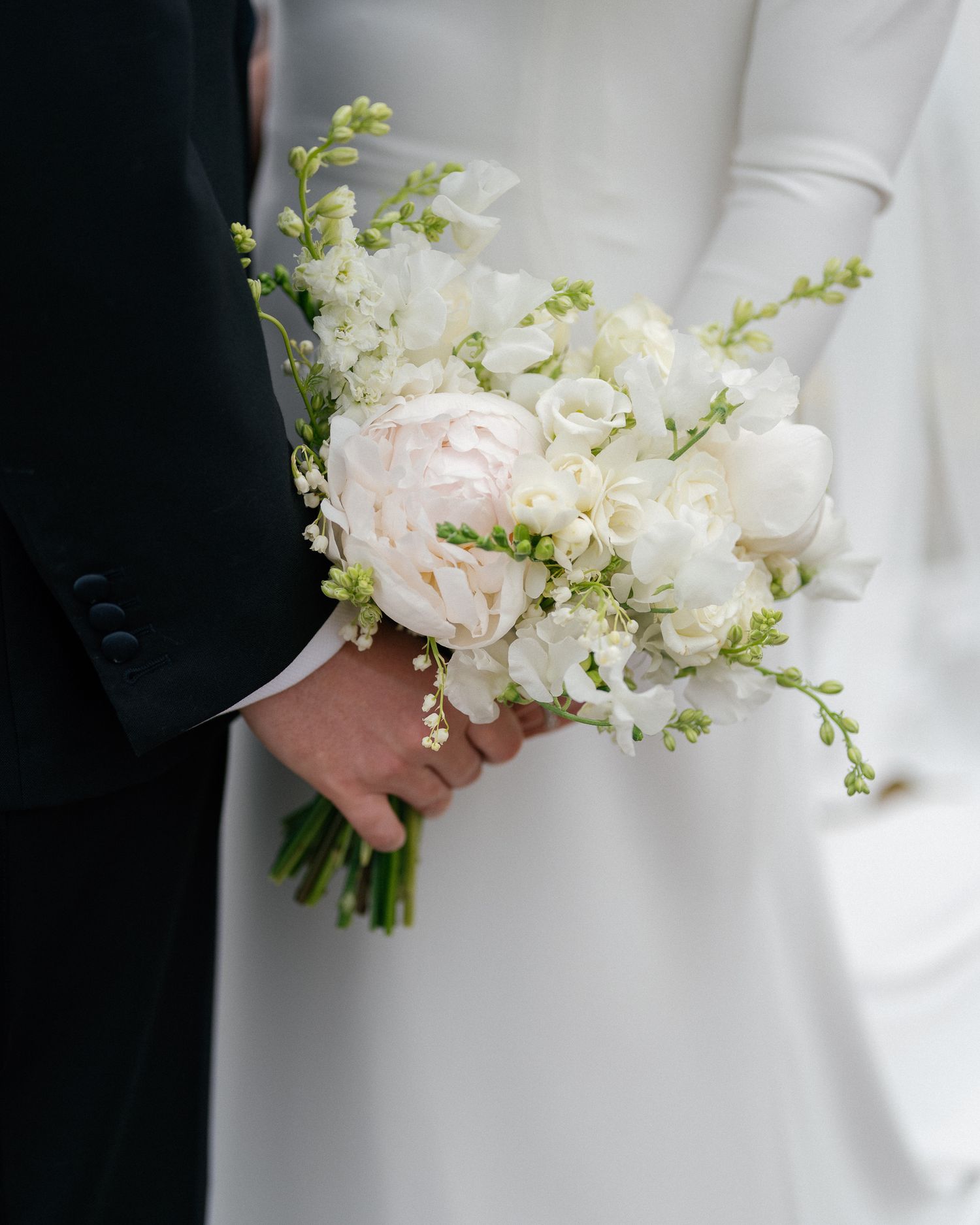 An elegant white and blush wedding bouquet with peonies and delicate green sprigs held against a white dress and dark suit.