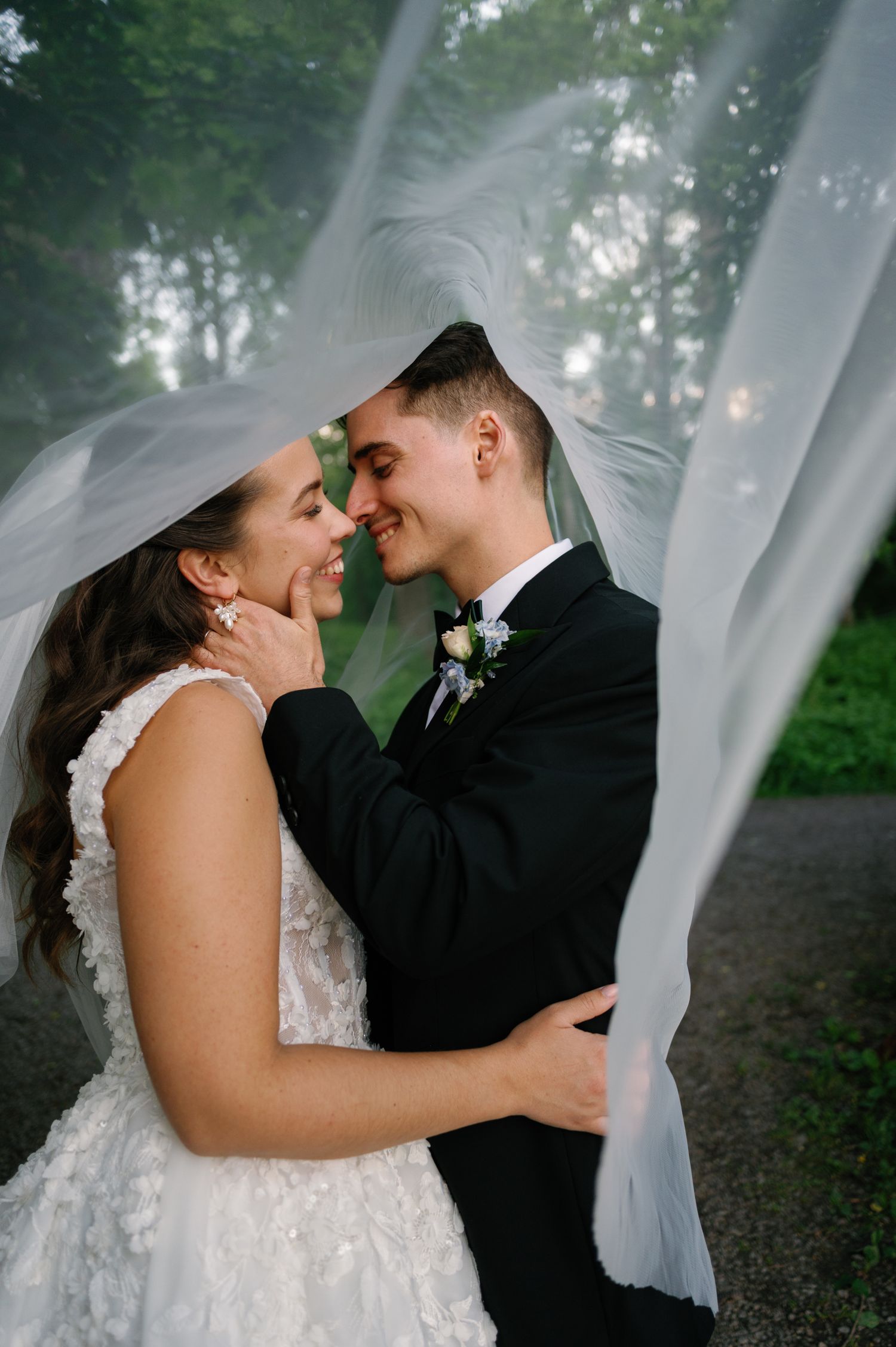 A romantic wedding couple embraces under a flowing white veil in a wooded setting.