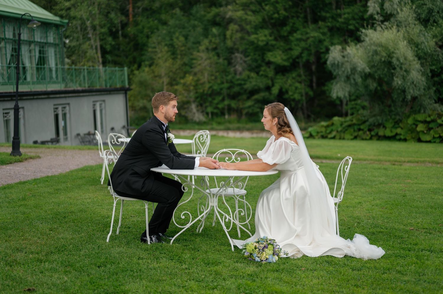 Wedding couple lounging on white garden furniture in an outdoor setting.