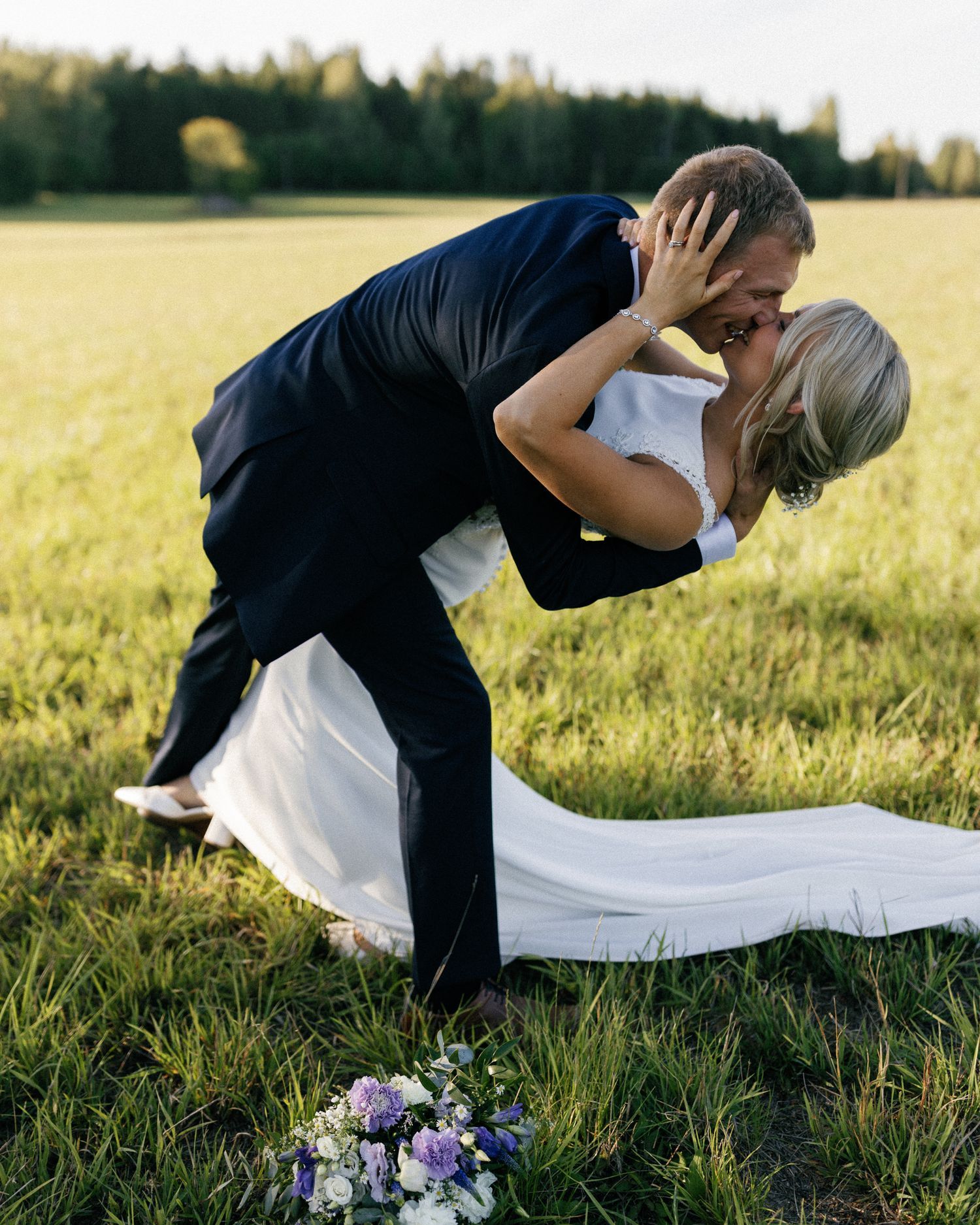 Dramatic dip kiss between newlyweds in sunny field with wedding bouquet on ground.