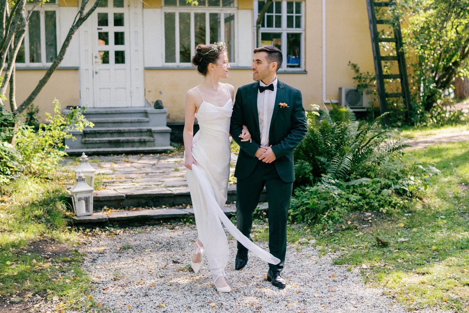 A couple walks together along a garden path outside a yellow house with white trim on a sunny day.