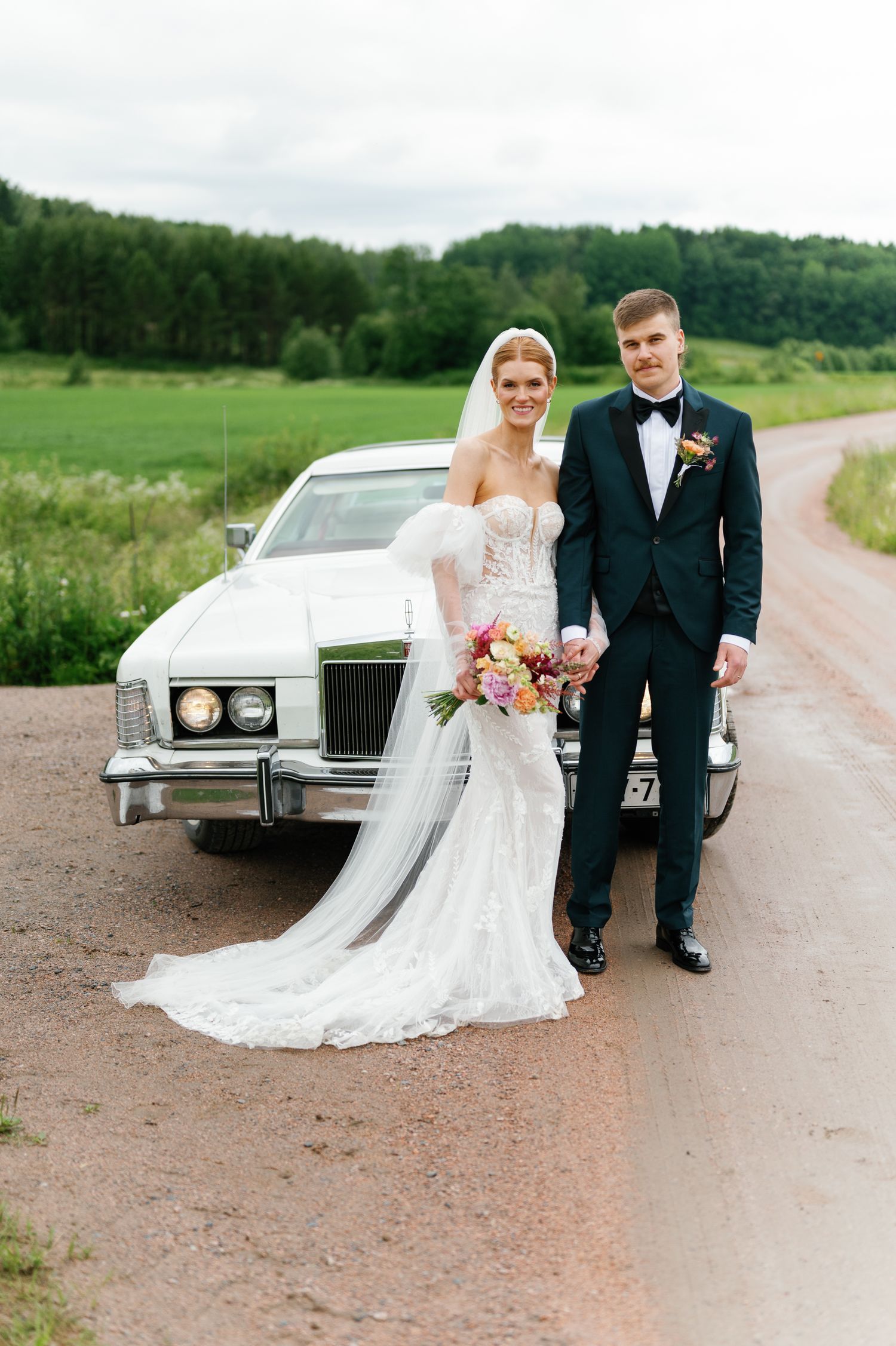 A wedding couple poses with a vintage white car on a country road.