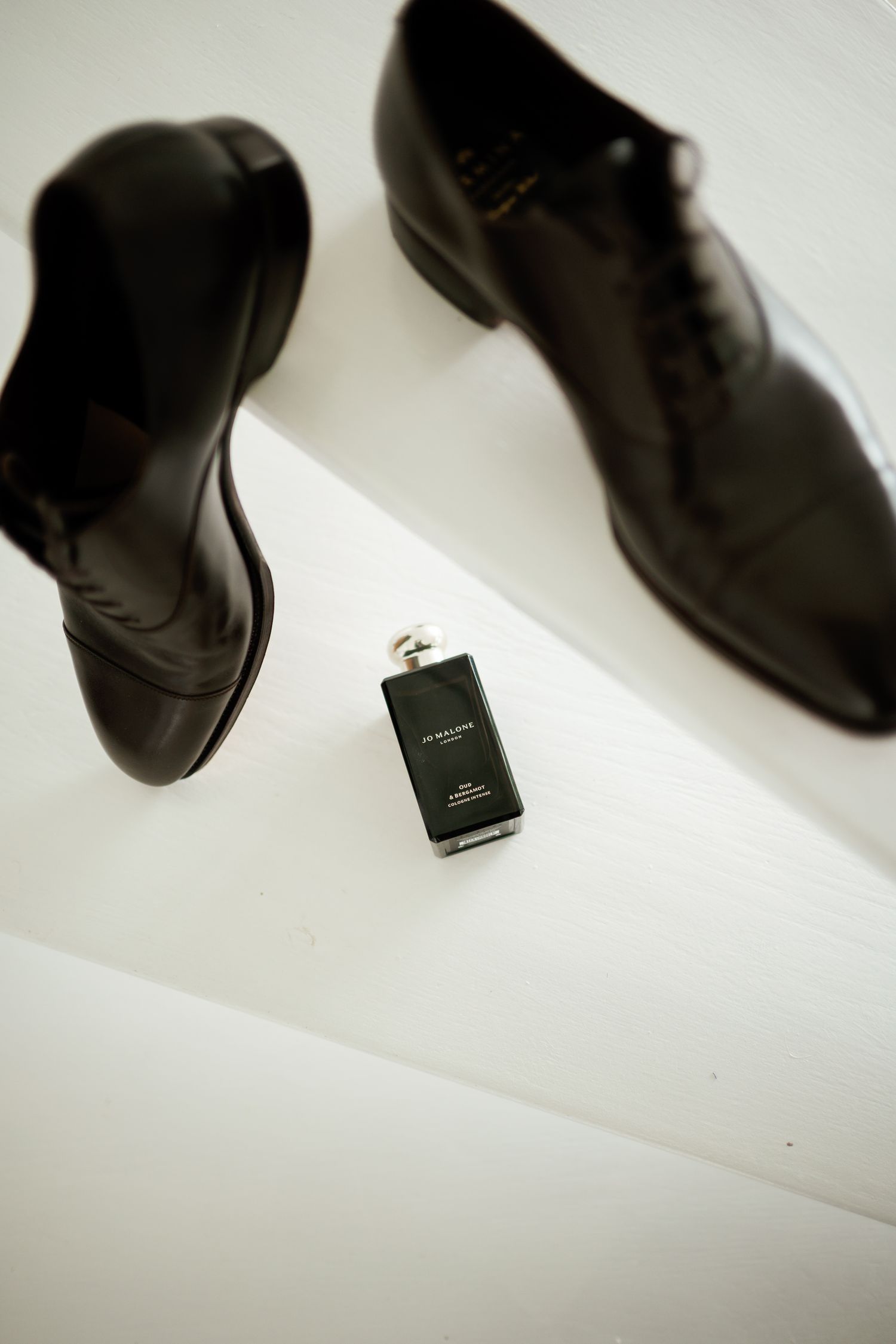 Black leather oxford dress shoes beside a small dark cologne bottle on a white surface.