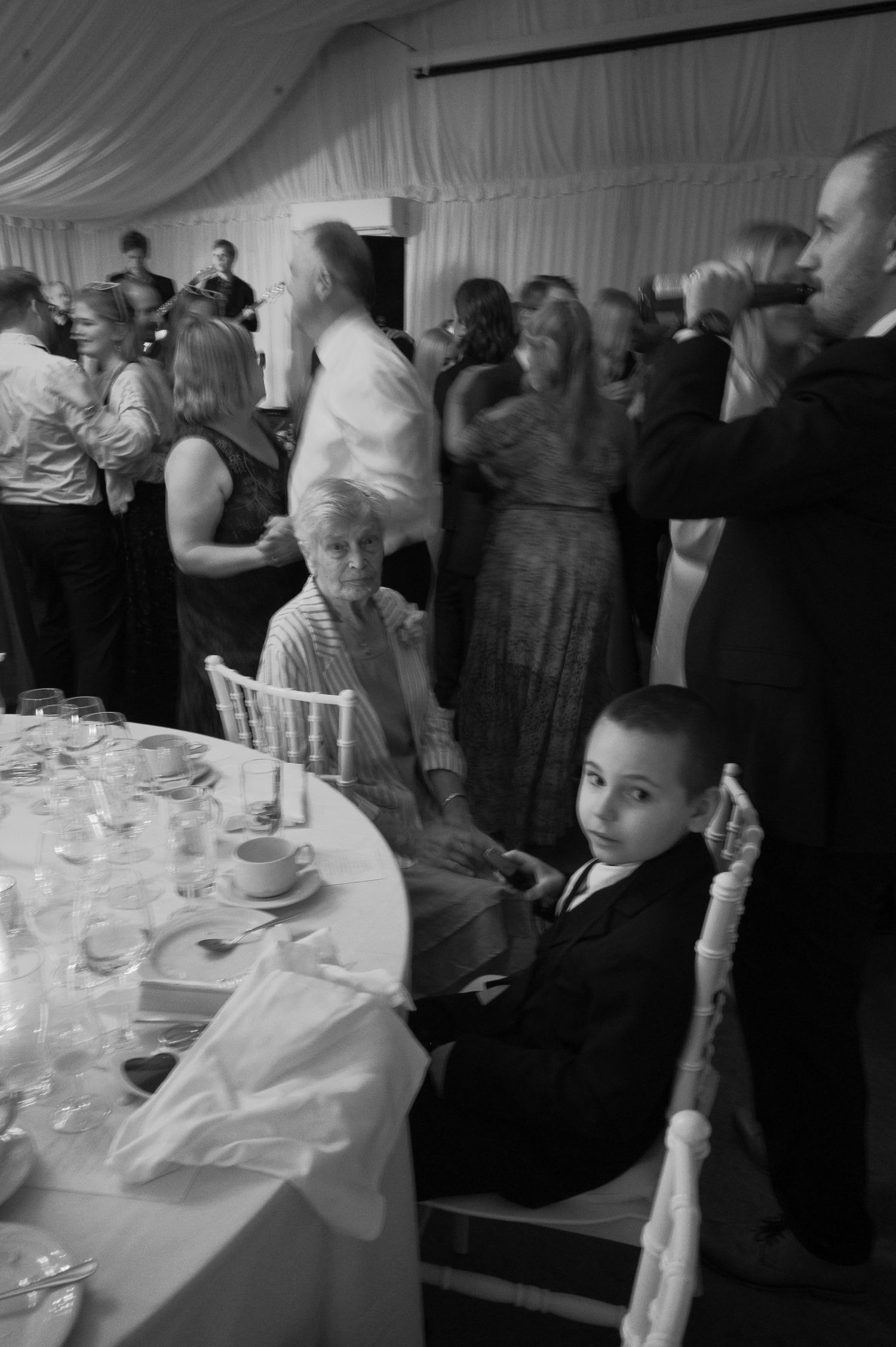Black and white photo of elegantly dressed guests at a formal event, featuring a young boy in a suit seated at a dining table.