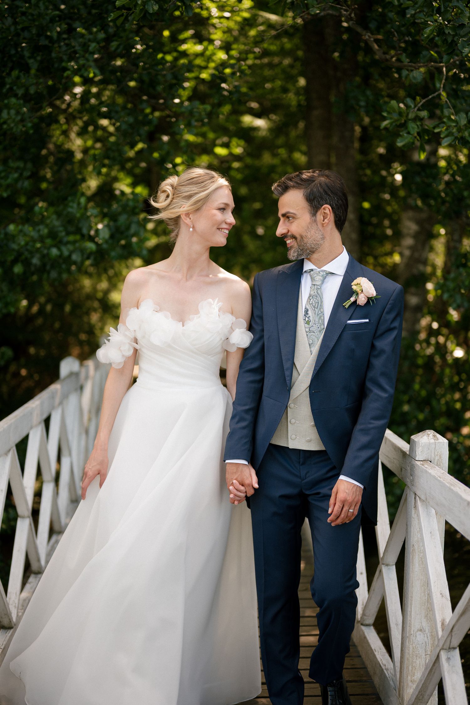 Bride and groom holding hands on a white wooden bridge, smiling at each other, surrounded by lush green trees.